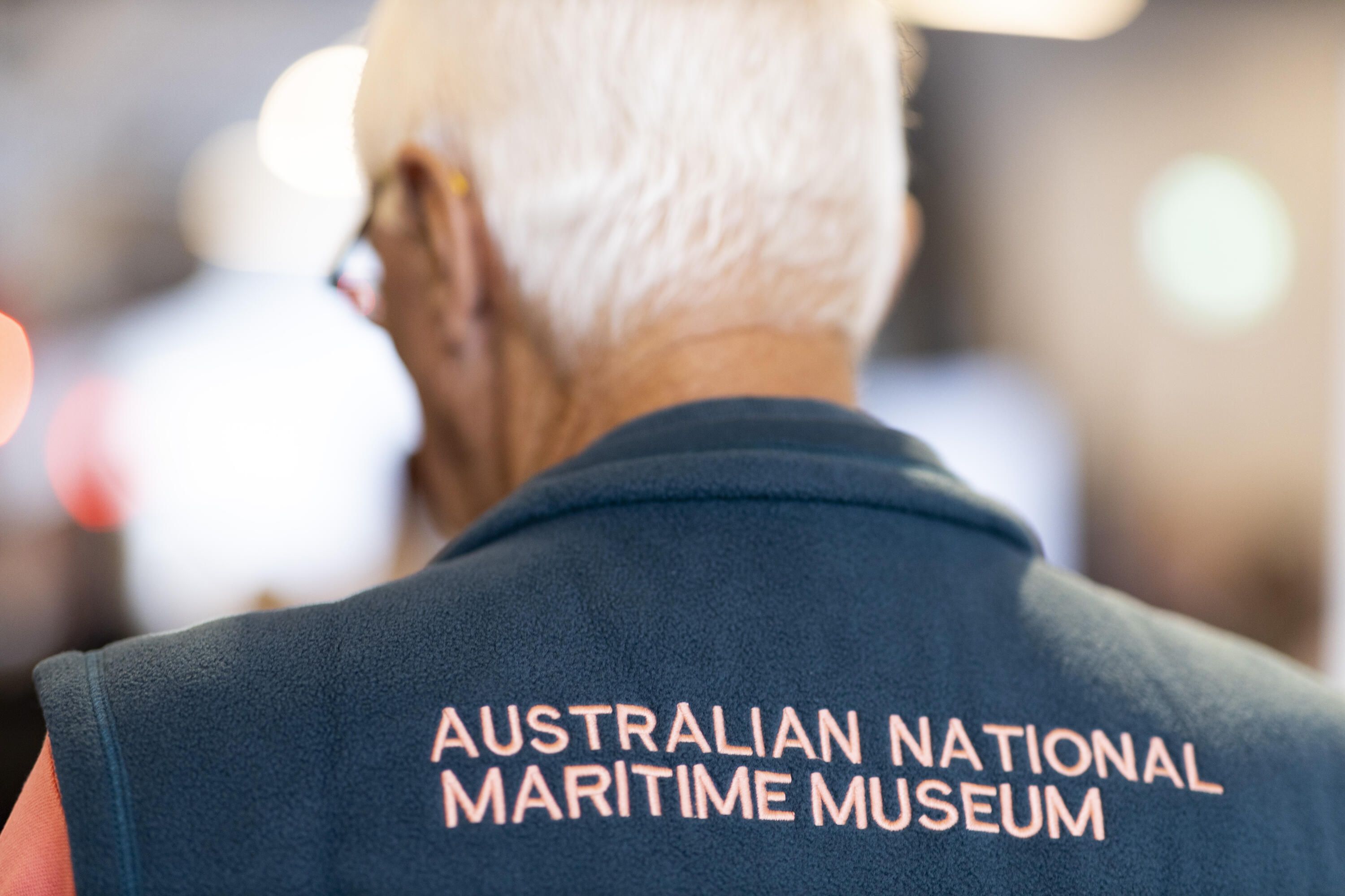 Back of a male museum volunteer with white hair wearing a vest with the text Australian National Maritime Museum.