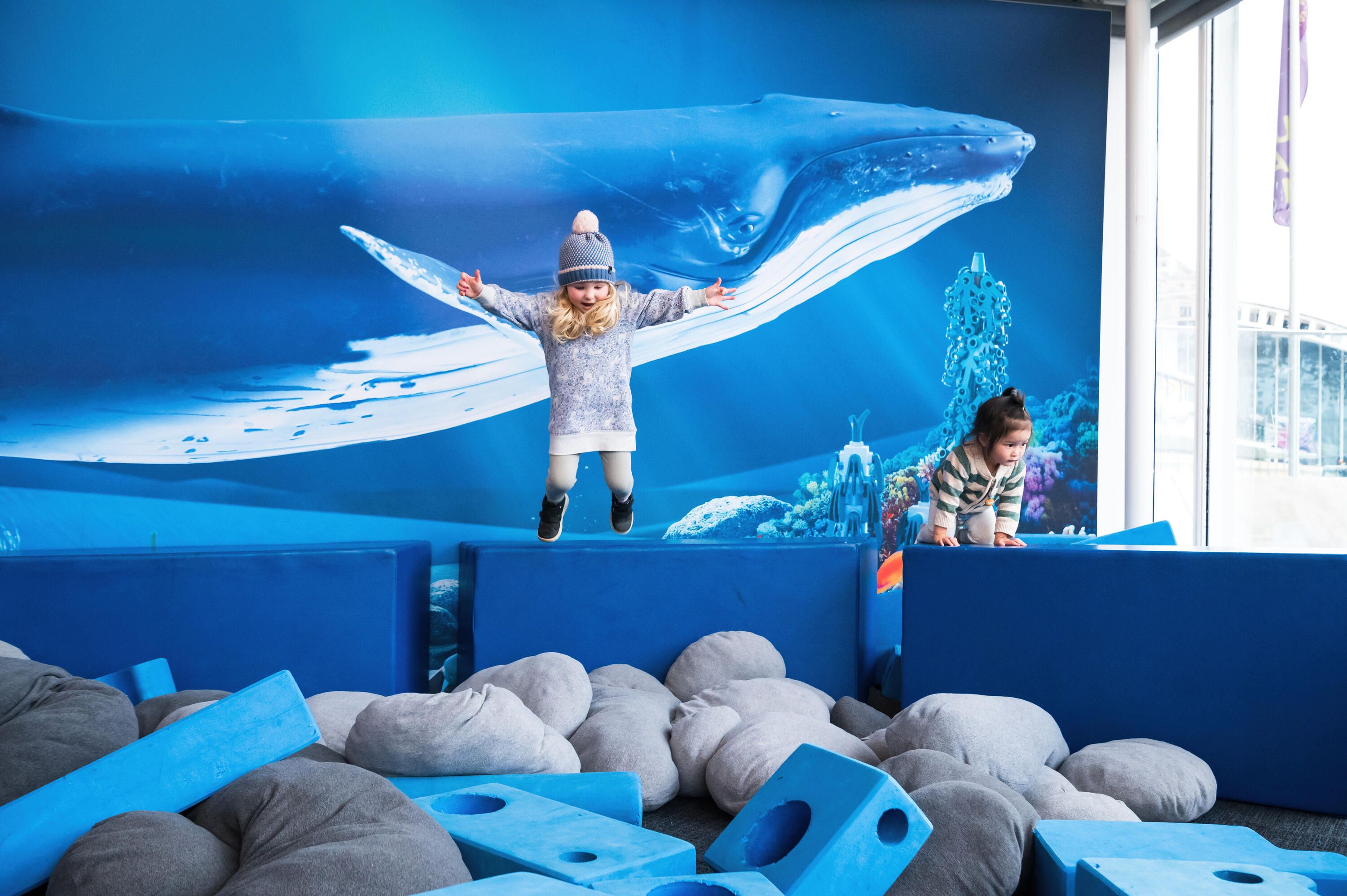 A small girl with blonde hair and a beanie jumping into a pile of grey cushions. 