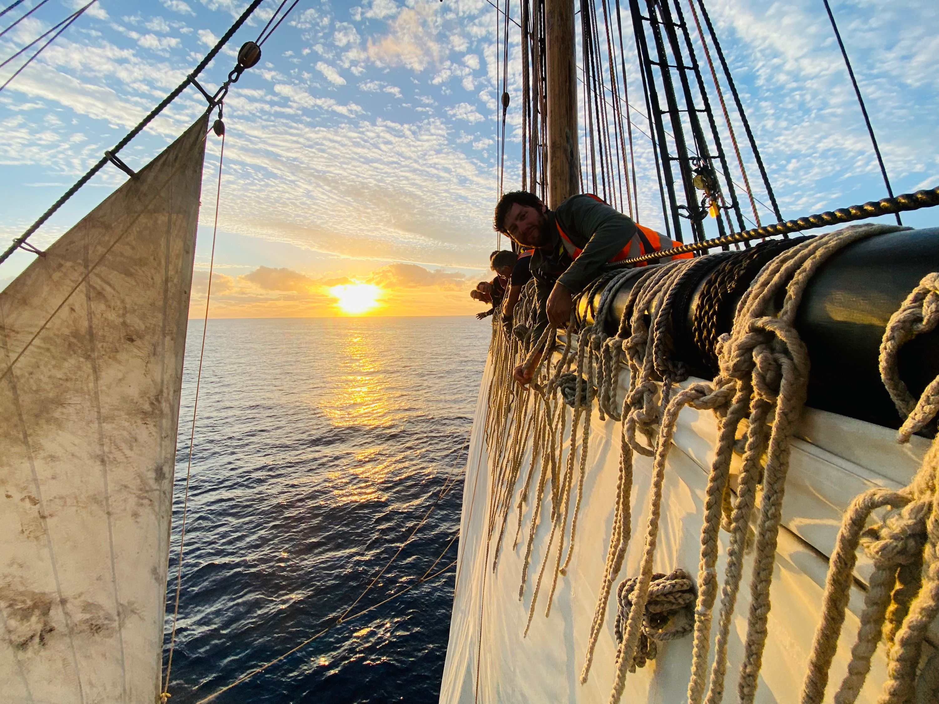 Photo up the rigging on a tall ship. It shows crew members spread along the yard working on the sails, with a sun on the horizon behind them.