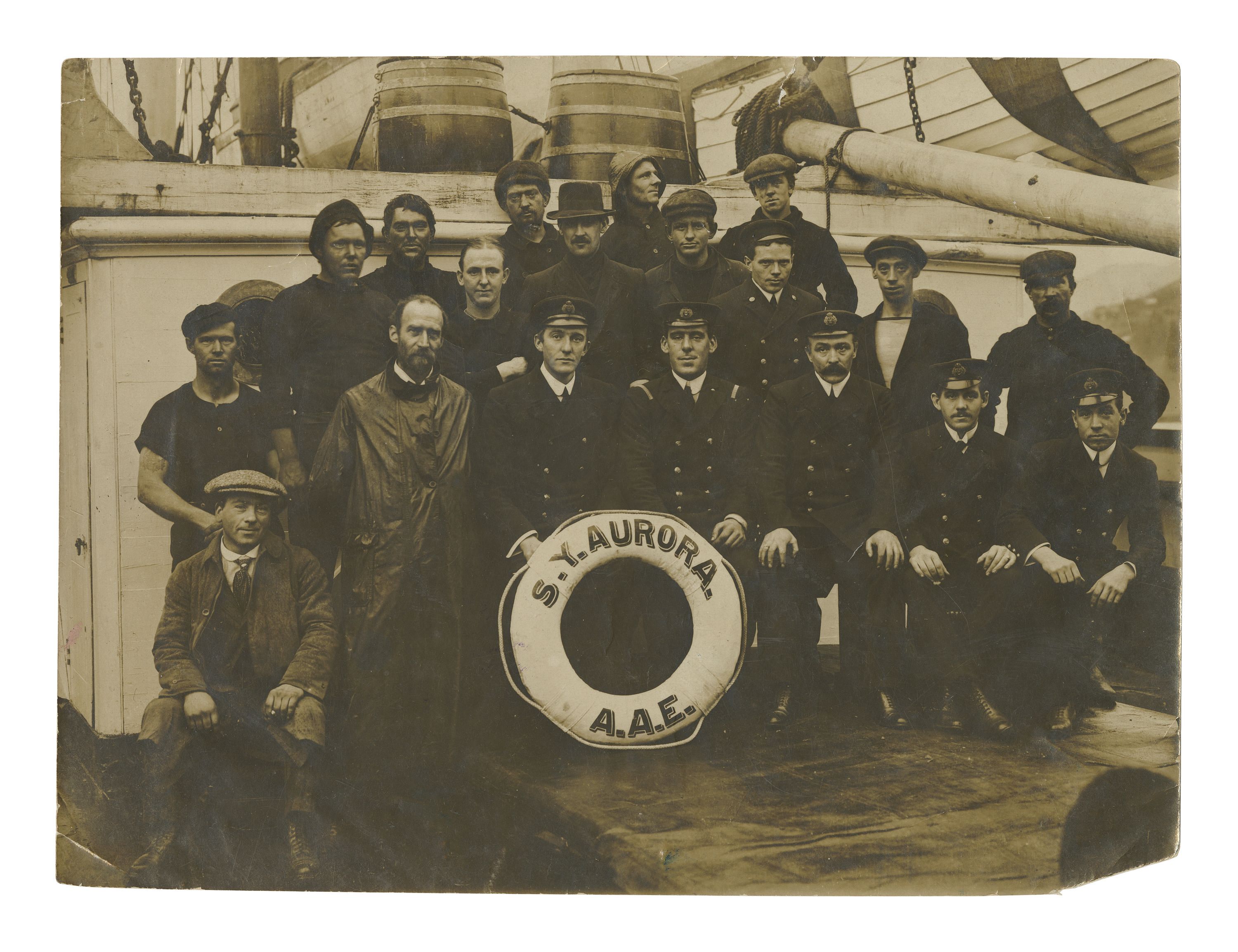 Aged, black and white photograph of members of the crew of the SY AURORA, a group of 19 men both seated and standing, in various clothing including formal uniforms, raincoats, and hats. In front of the group is one of the vessel's white life rings, featuring the text: SY AURORA 