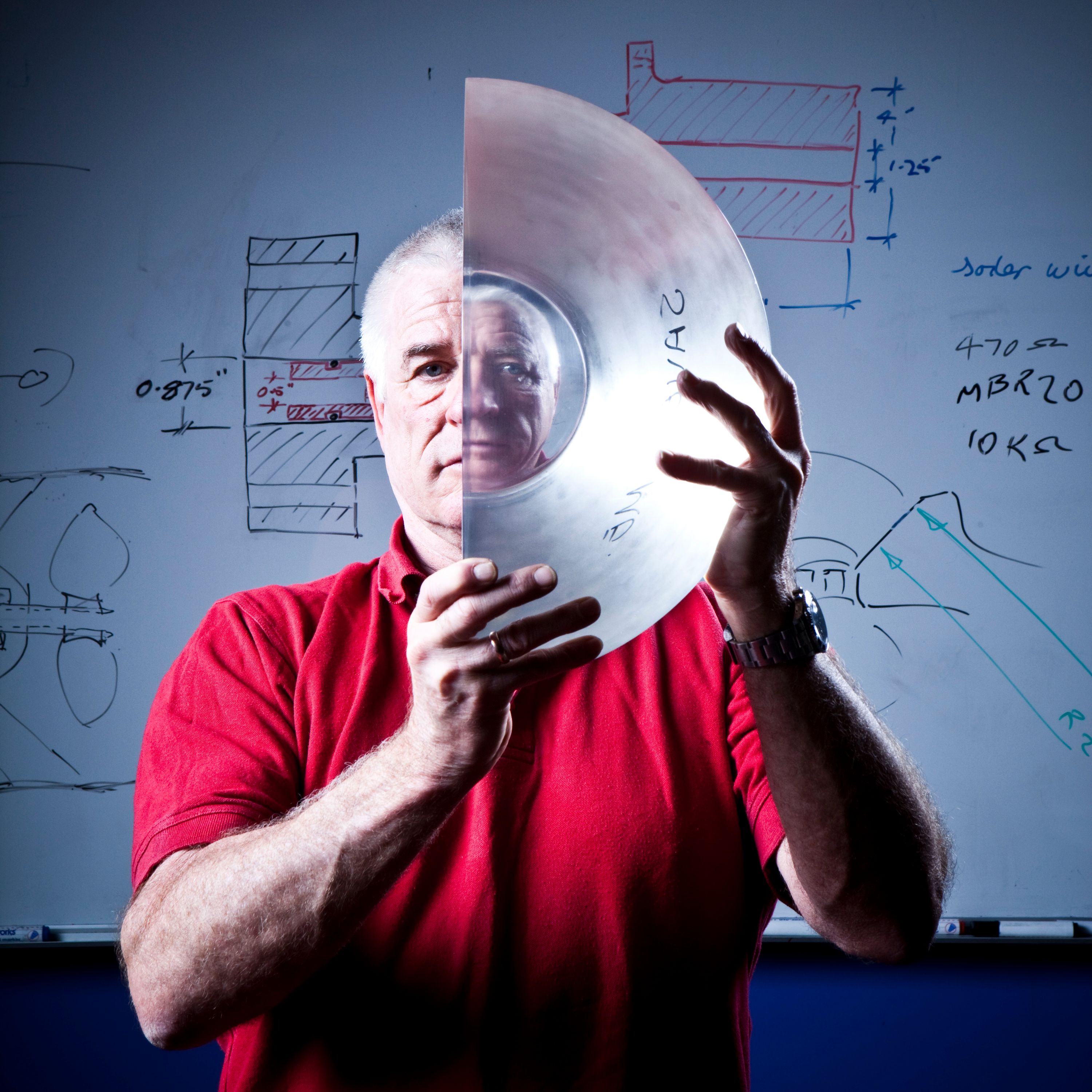 A man with white hair and a red shirt is looking towards the camera, holding a half circle lenze, making the right half of his face appear smaller.