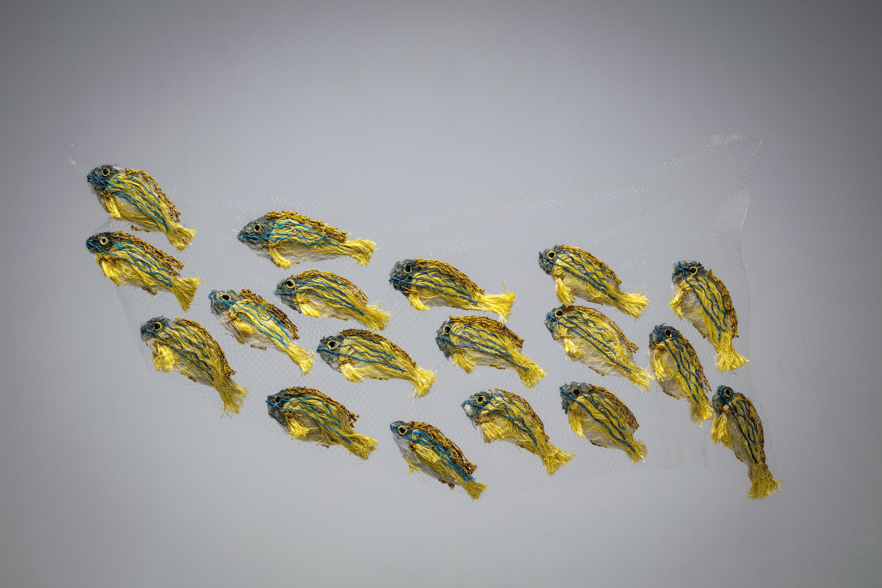 Sculpture of a school of golden snapper fish with blue stripes. The backing is a clear net, with the snapper being predominately yellow in colour with four blue stripes. Each fish has a grey and white face and a white belly.