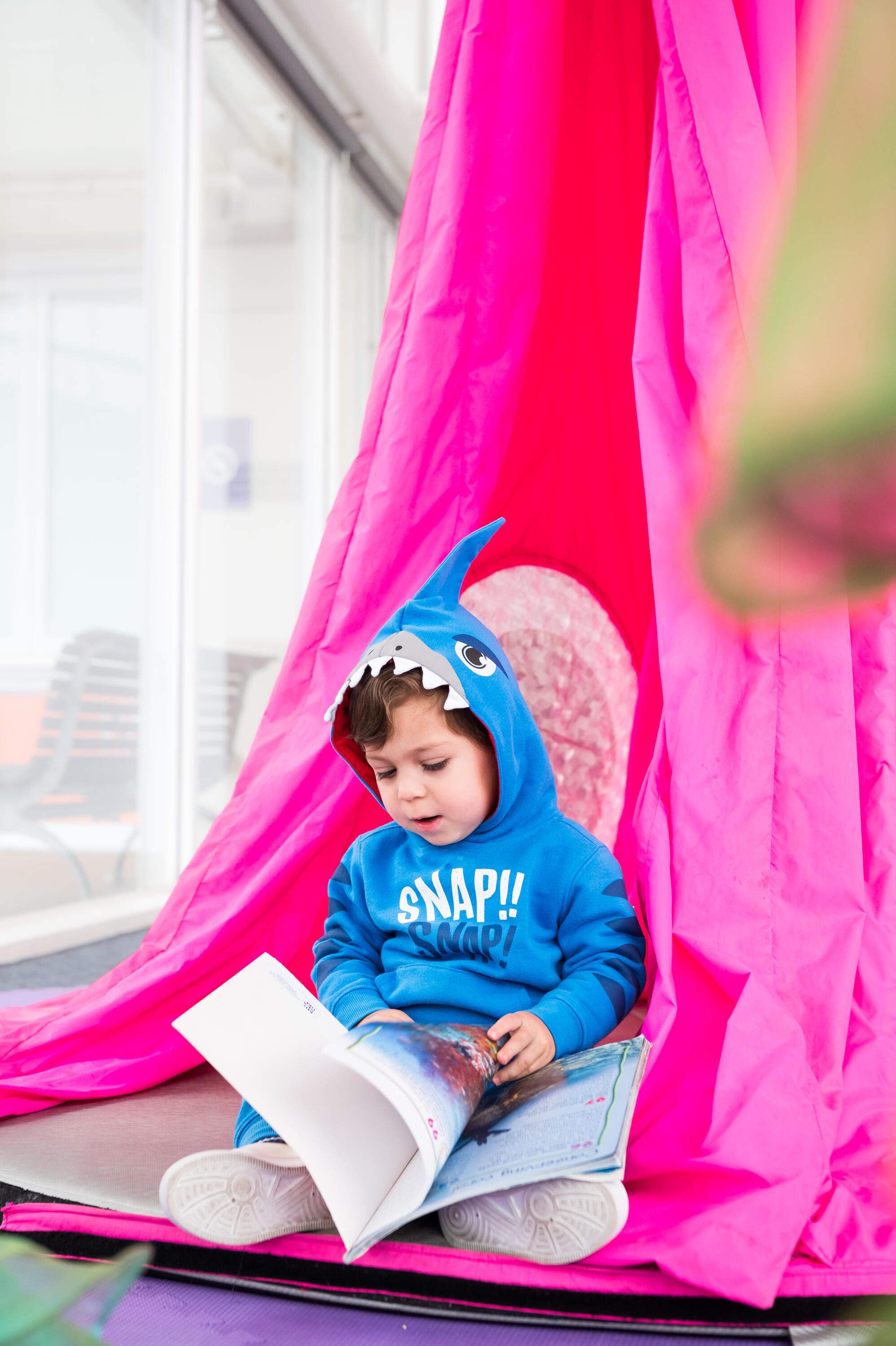 Photo of smiling boy wearing a blue hoodie with shark teeth, siting in front of a bright pink tent, reading a book.