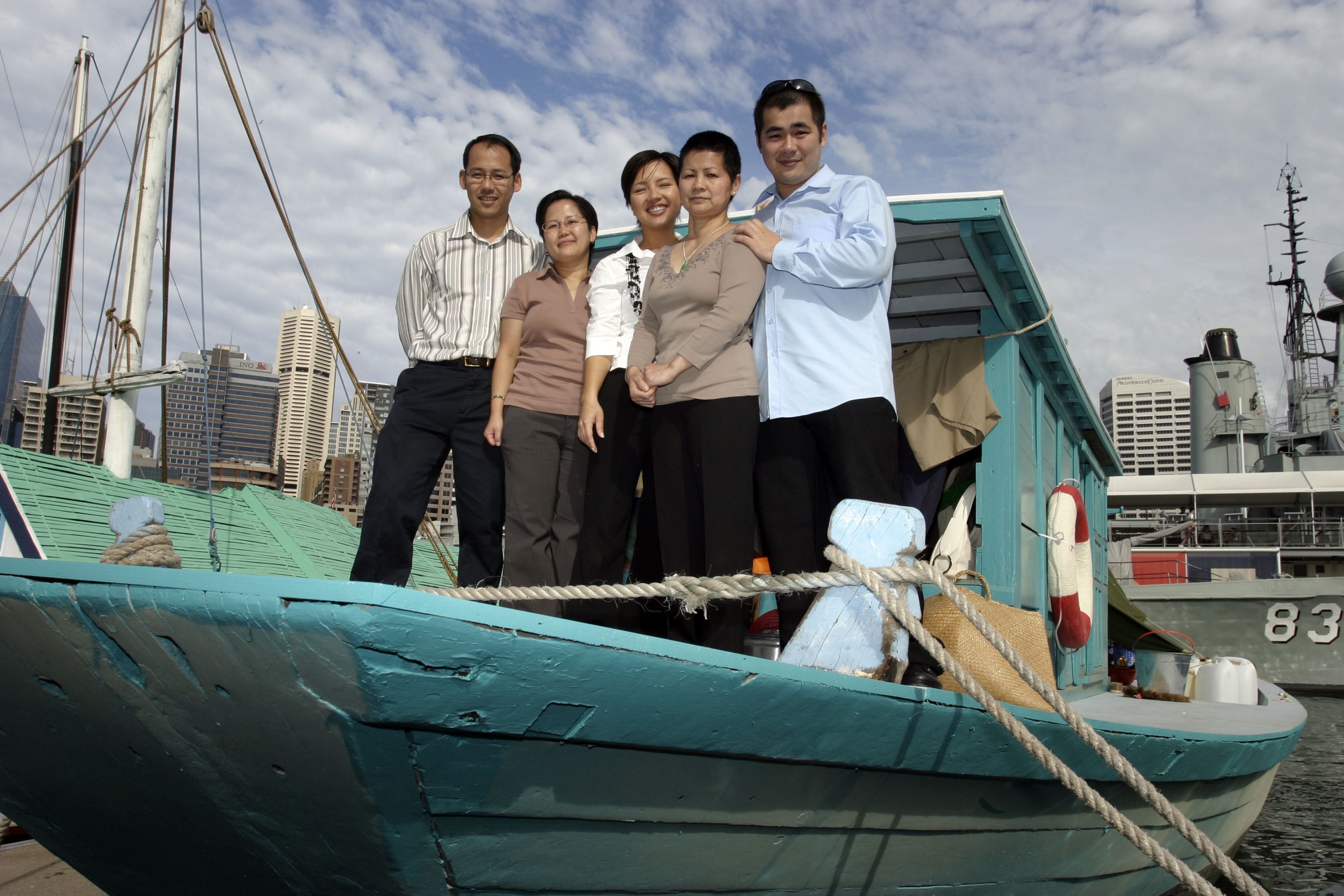 Photograph showing a family, with 5 adults standing on the deck of a wooden boat which is painted blue. 