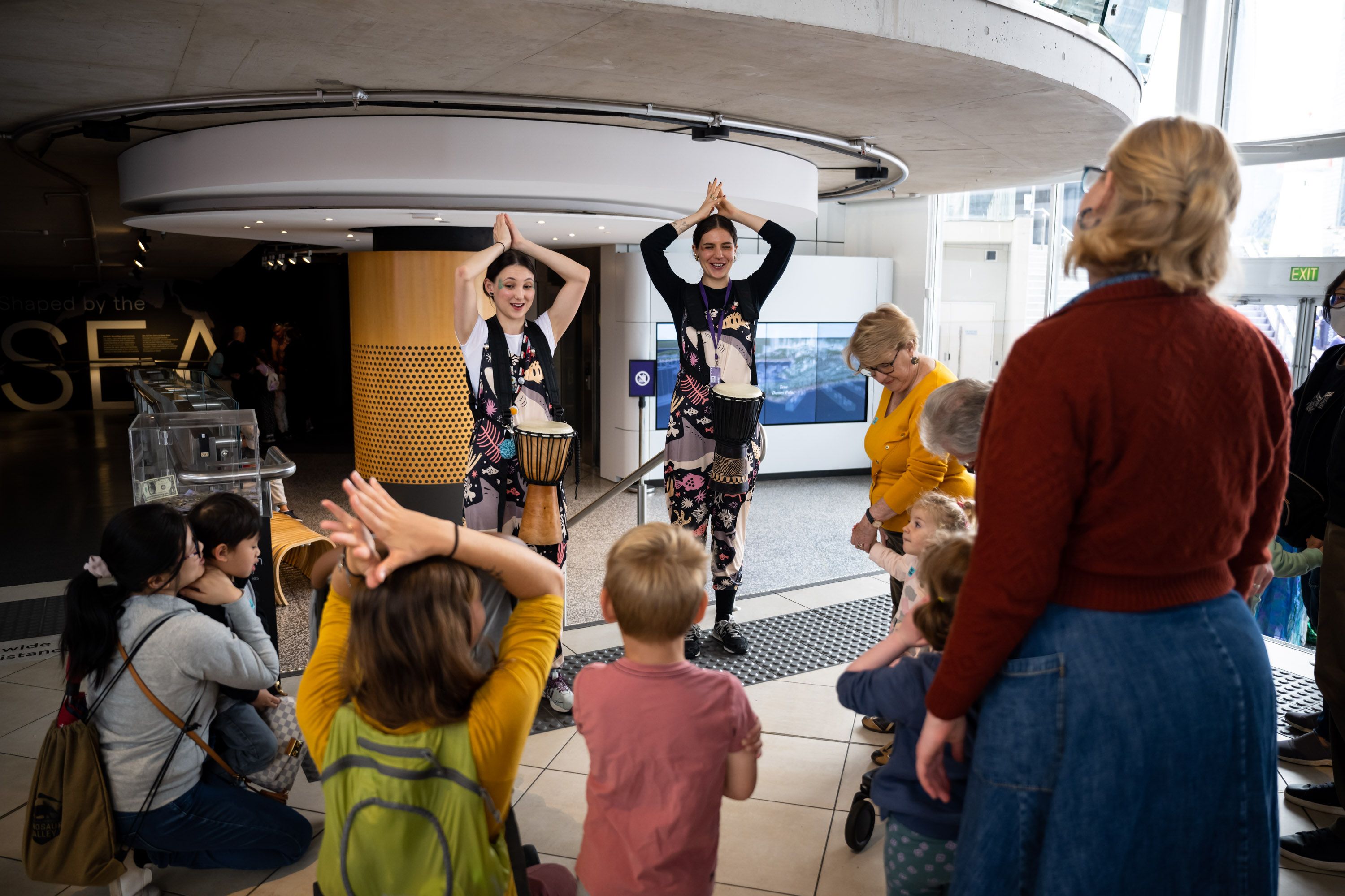 Two museum staff wearing costumes and drums are doing actions, surrounded by a group of young children and their parents