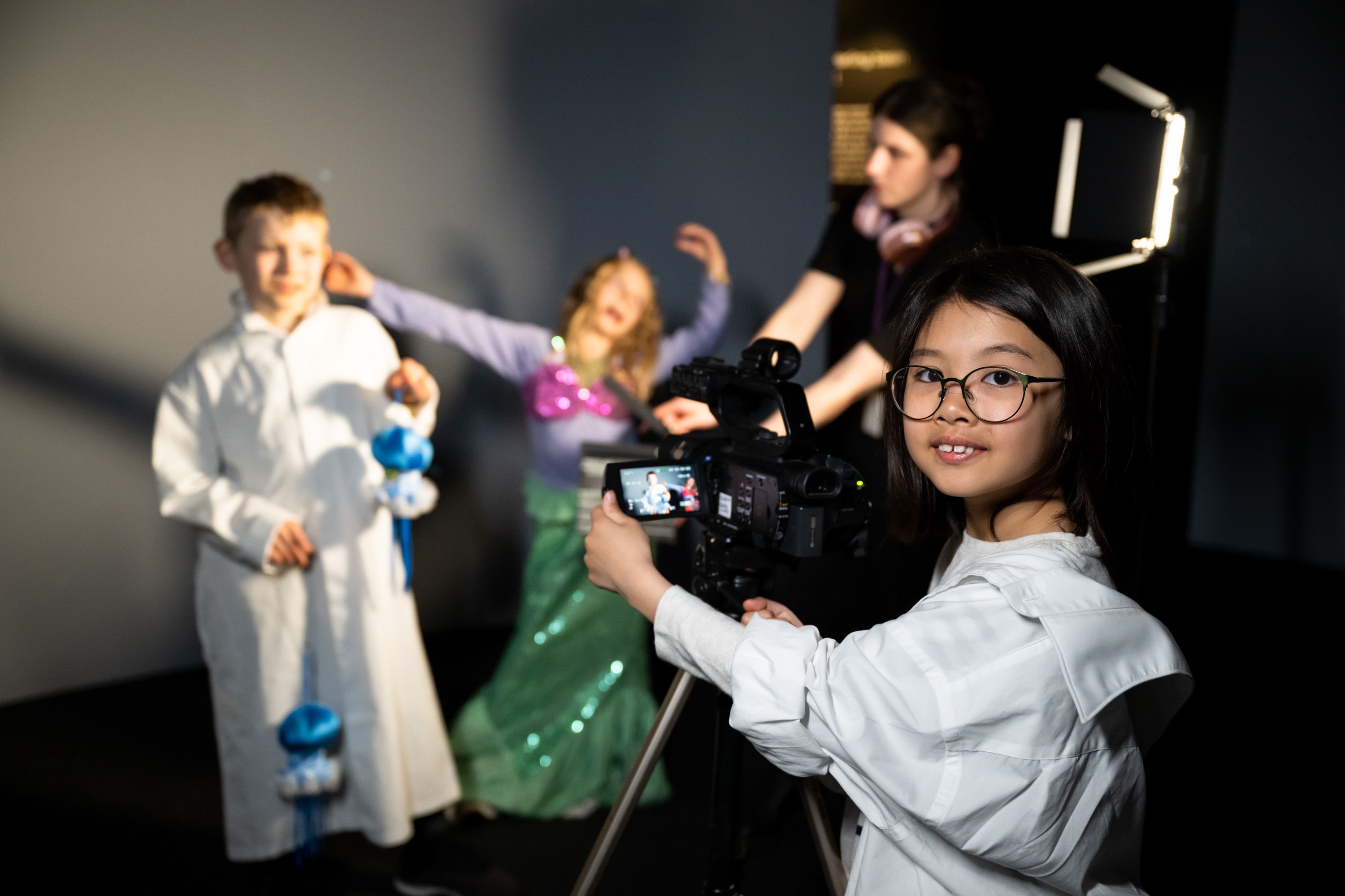 Photograph of a young girl with black hair and glasses, operating a camera on a tripod pointing towards 2 other children in the background.