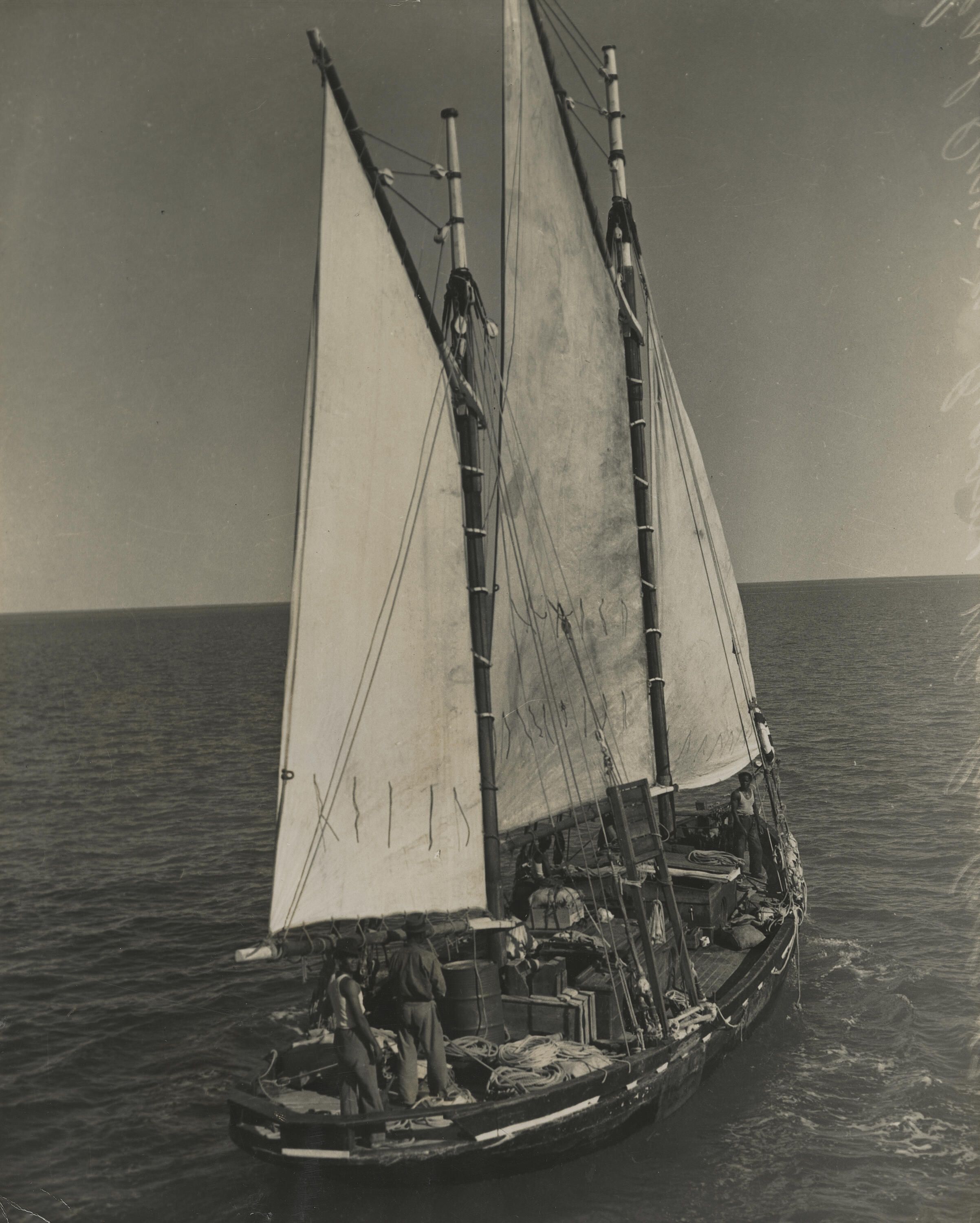 lack and white photograph on yellowed paper showing a pearling lugger boat with full sail. several people canbe seen onboard