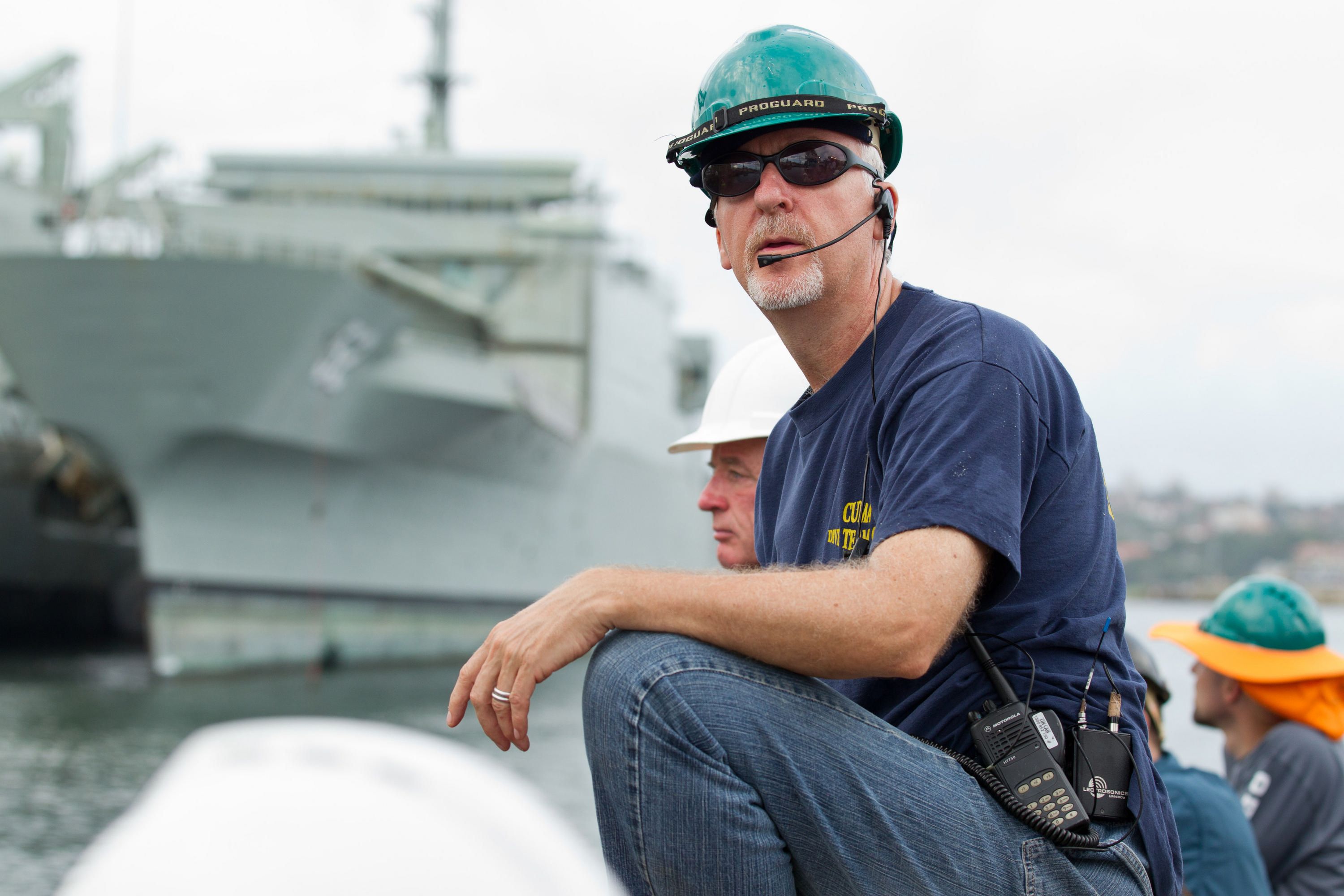 Photo of a man, James Cameron, wearing a helmet and radio, blue t-shirt and jeans. There is a grey ship in the background.