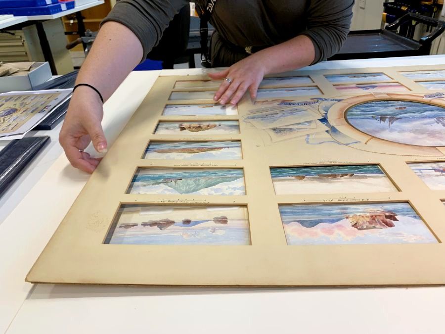Photo showing a person carefully adjusting a mount with many windows, each containing a watercolour.