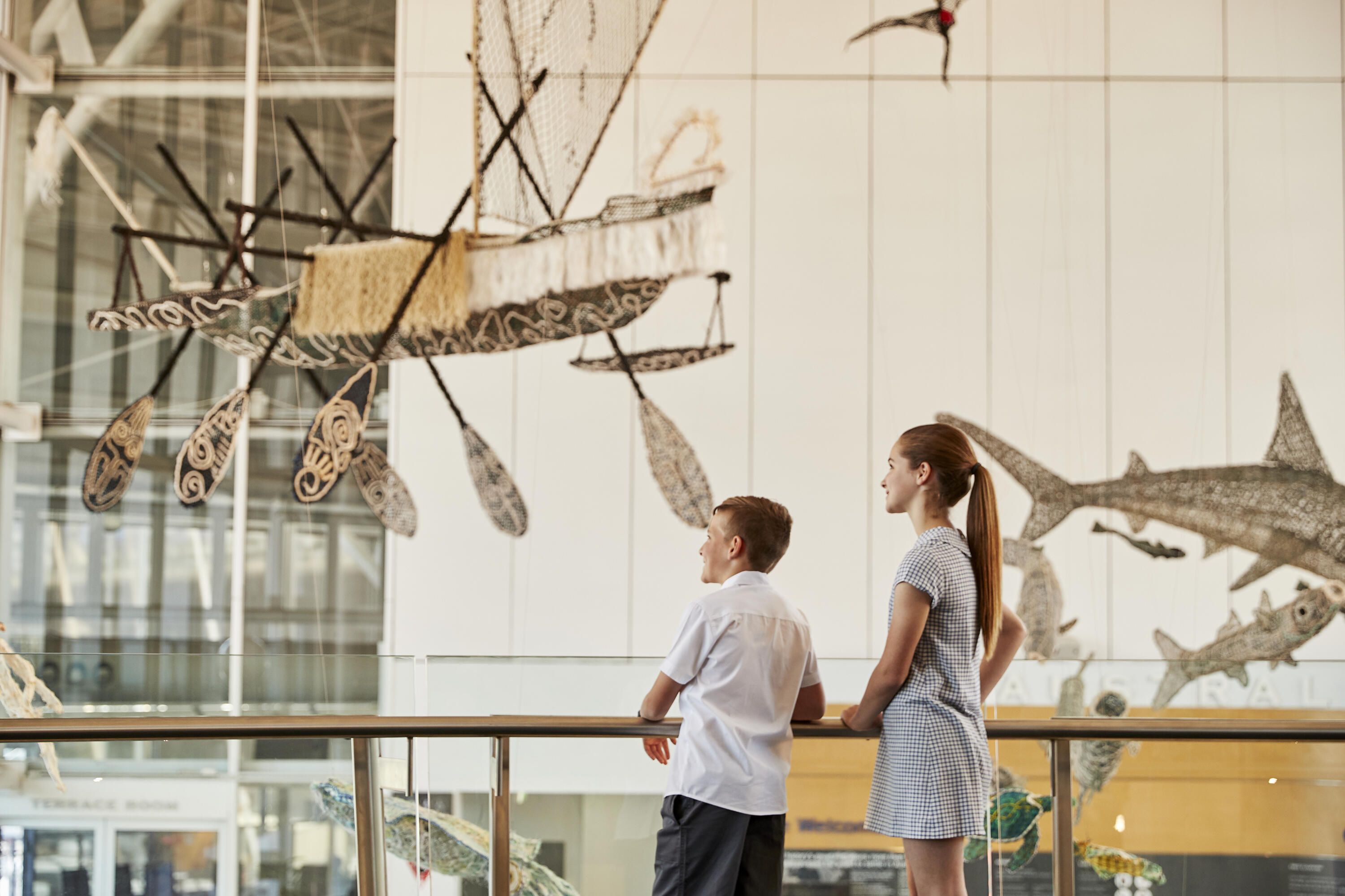 Photo of school kids exploring the museum, stop to look at the ghost net sculptures suspended in the foyer.