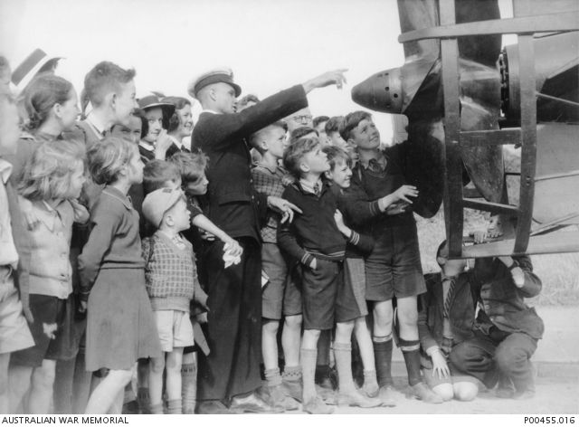 Black and white photo showing a man in a dark, formal uniform pointing at an object, the rear propeller of a submarine. He is surrounded by a crows of people, mostly children in school uniforms.