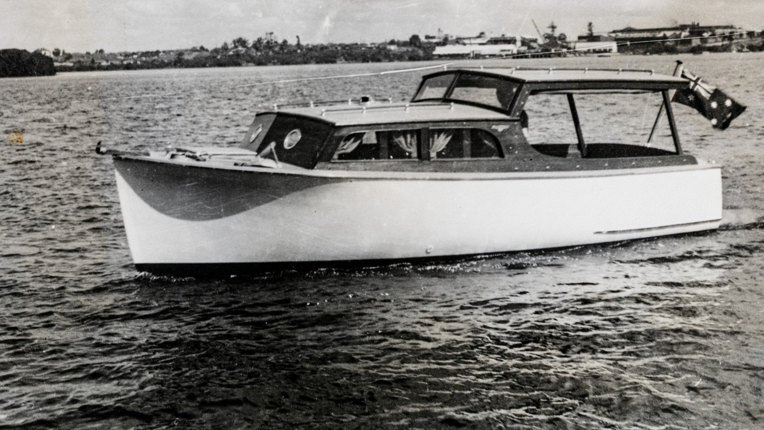 black and white photo of a motor cruiser with a white hull and an Australian flag flying off the rear. 
