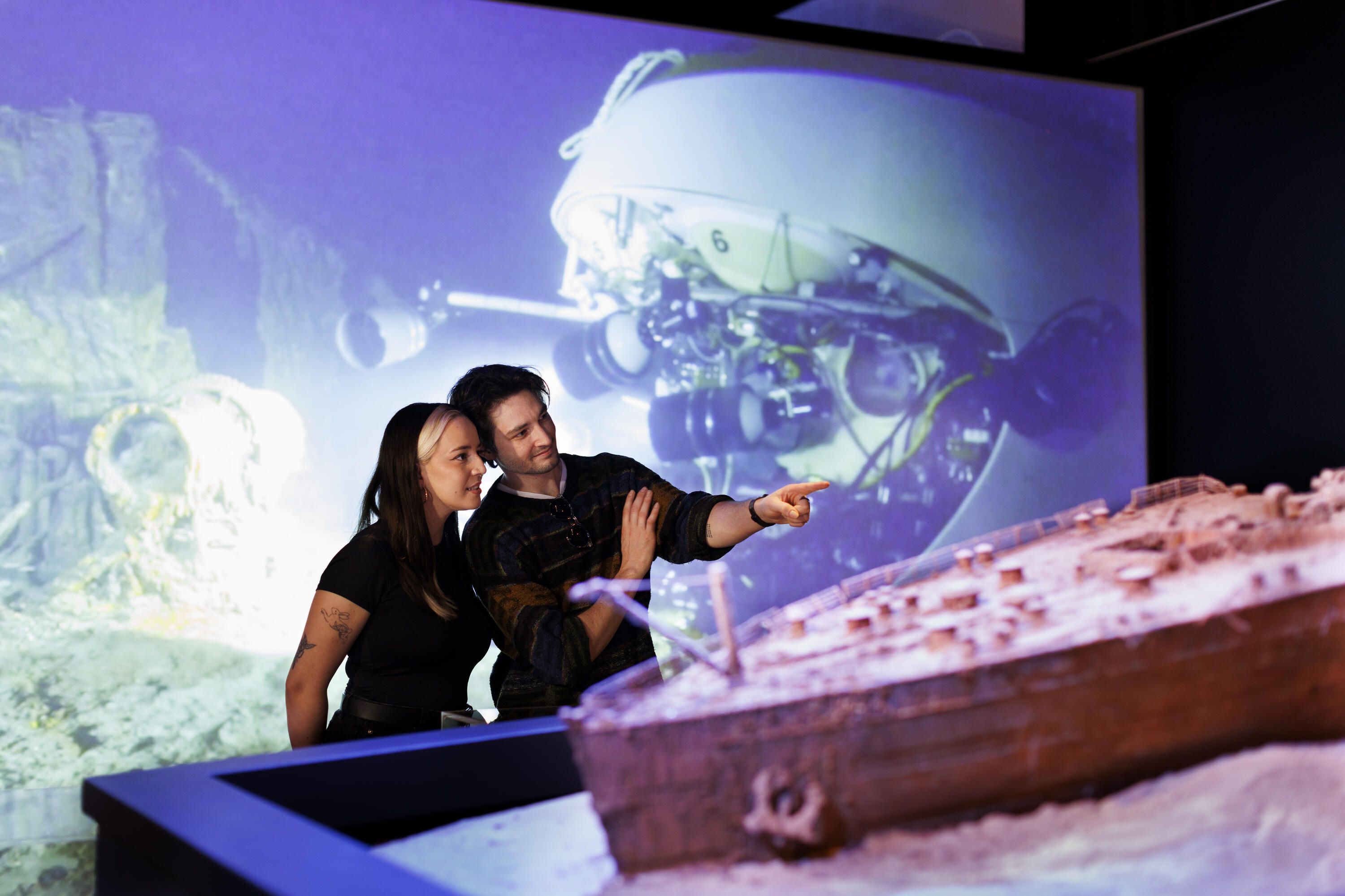 Two people, a man and a woman point at a ship model which is out of focus in the foreground, with a large projection of underwater maritime archaeology behind them.  