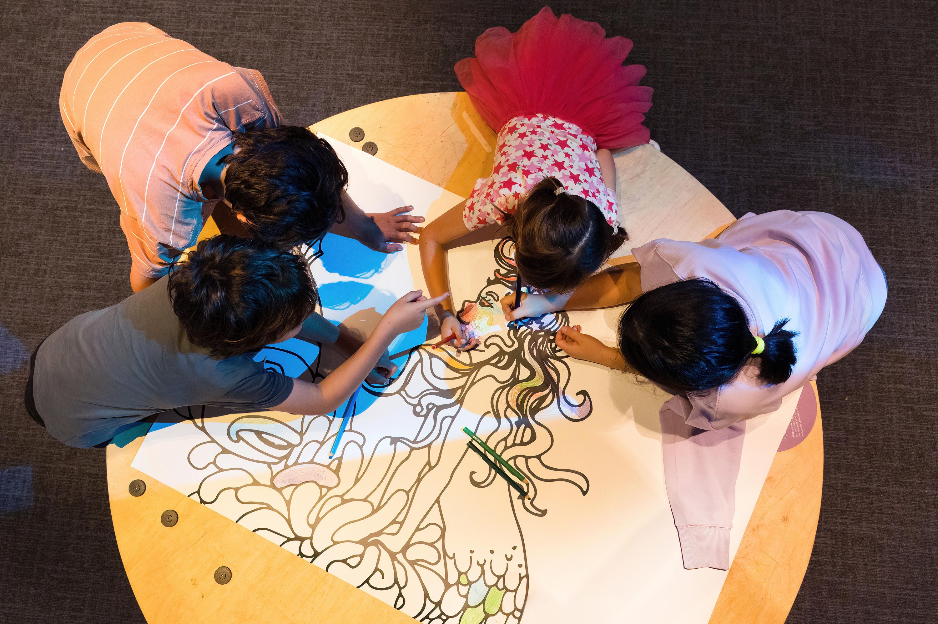Photo taken above a group of children participating in a collaborative colouring activity inside an exhibition space.