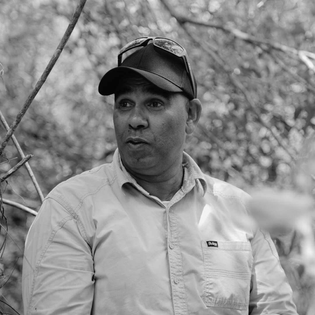 Black and white photo of a man with. darker skin, cap and button down shirt with trees in the background
