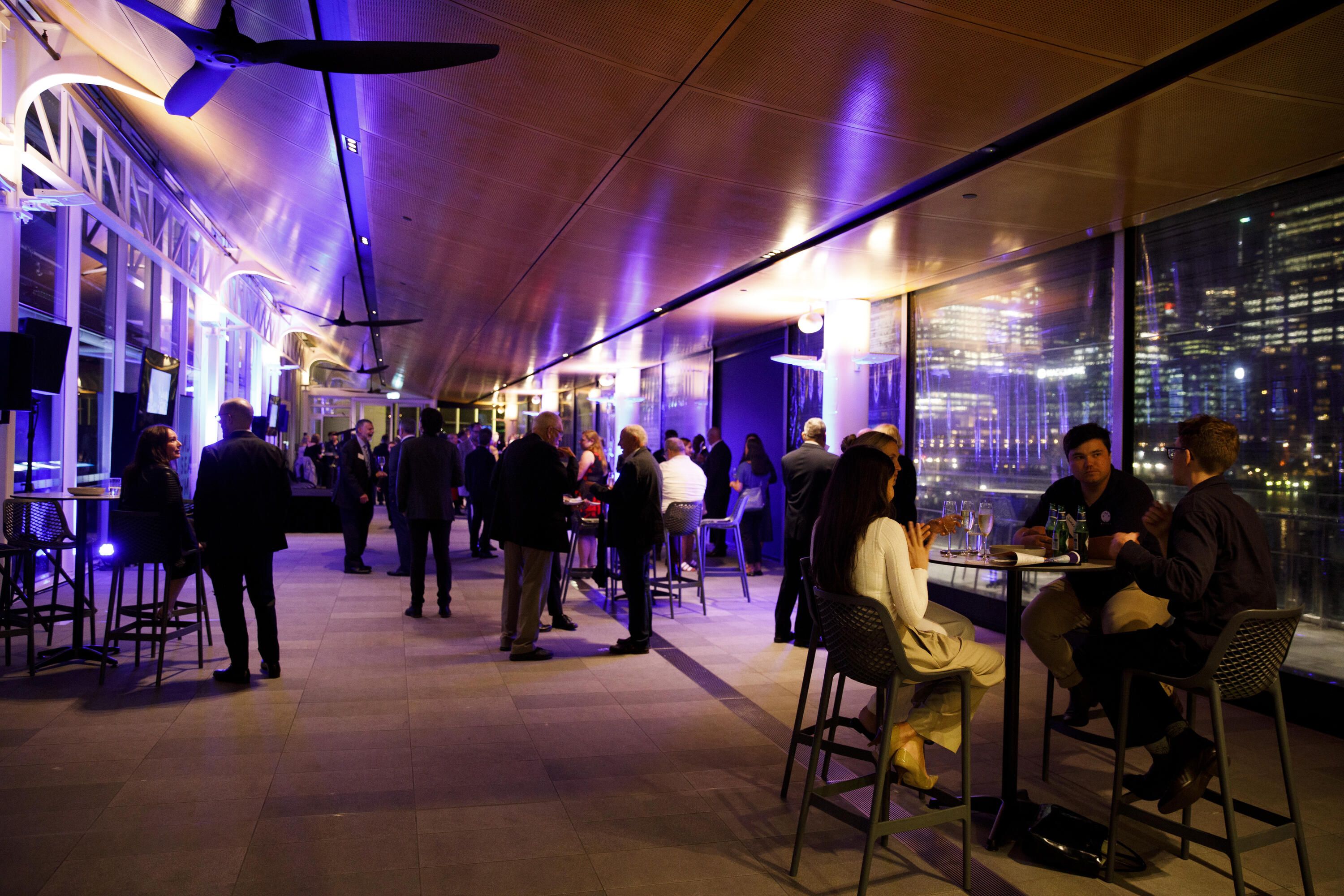 Photo showing guests at an event. It it night with colourful lighting and a city view in the background.