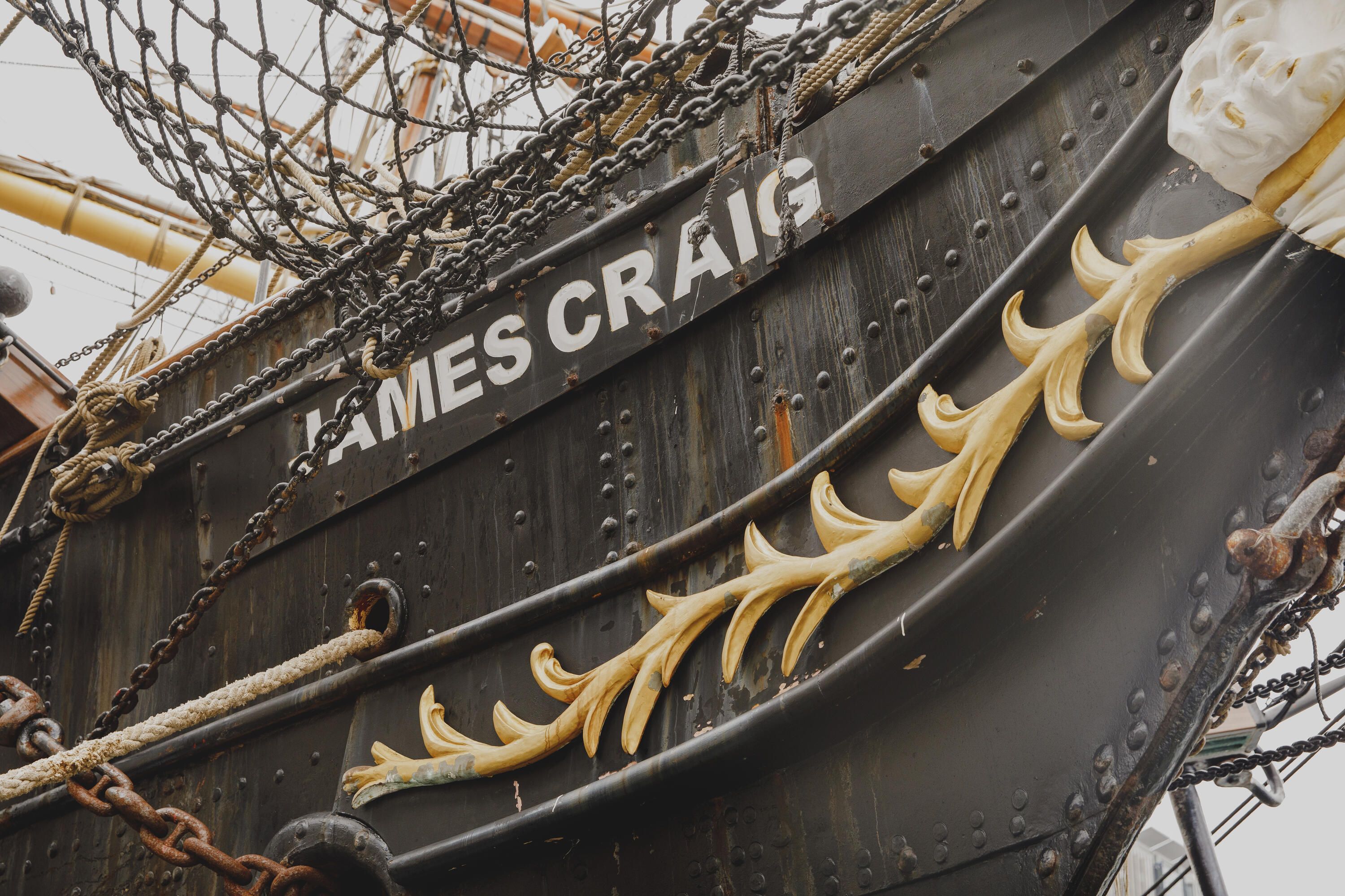 Close up photograph showing the front of a tall ship, with the name "James Craig" painted on the black hull. There are also ropes and gold details.