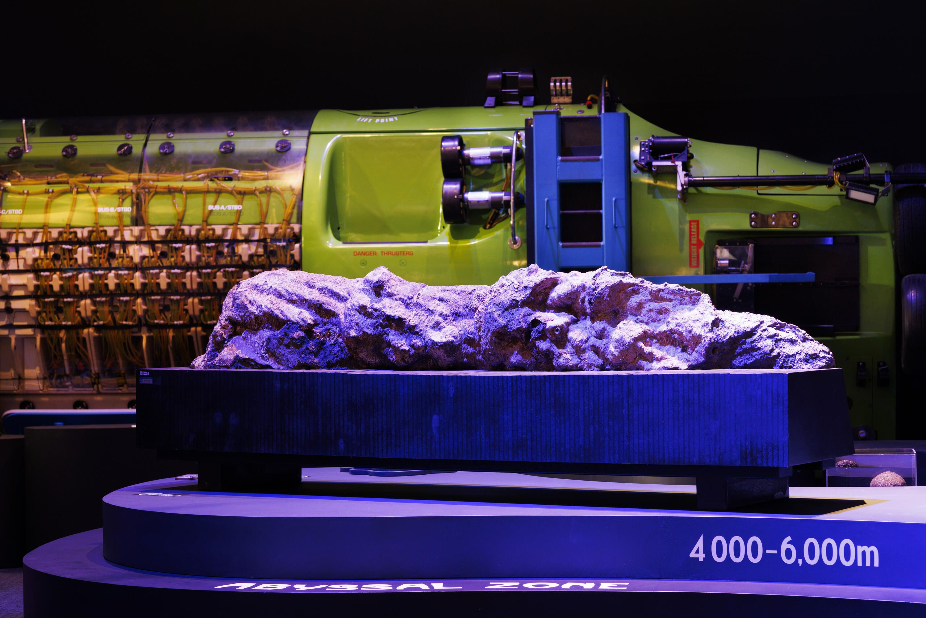 Photo showing a long, rock like object displayed on a plinth, behind it is part of a lime green submarine.