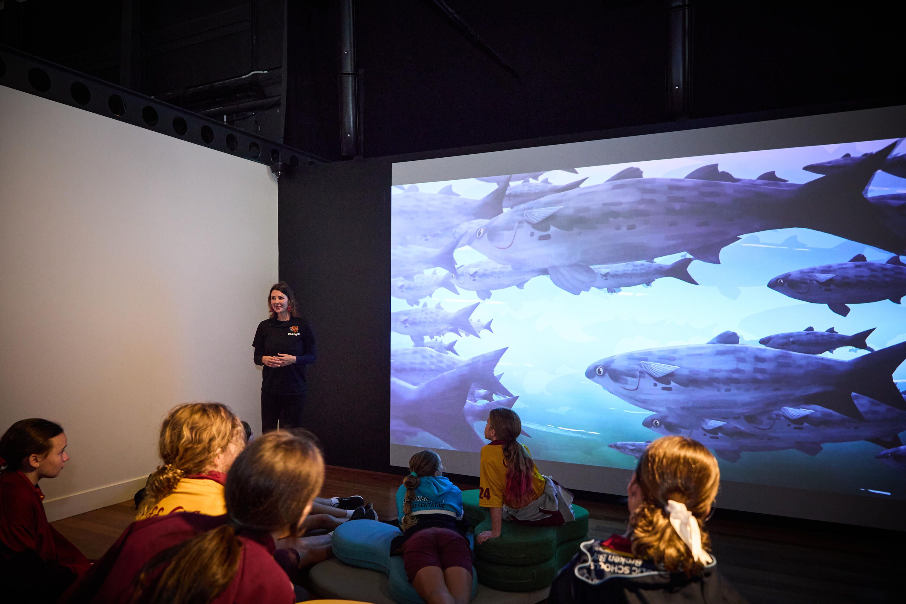 Photo of students in a museum exhibition space with curved soft seating and a large projection screen