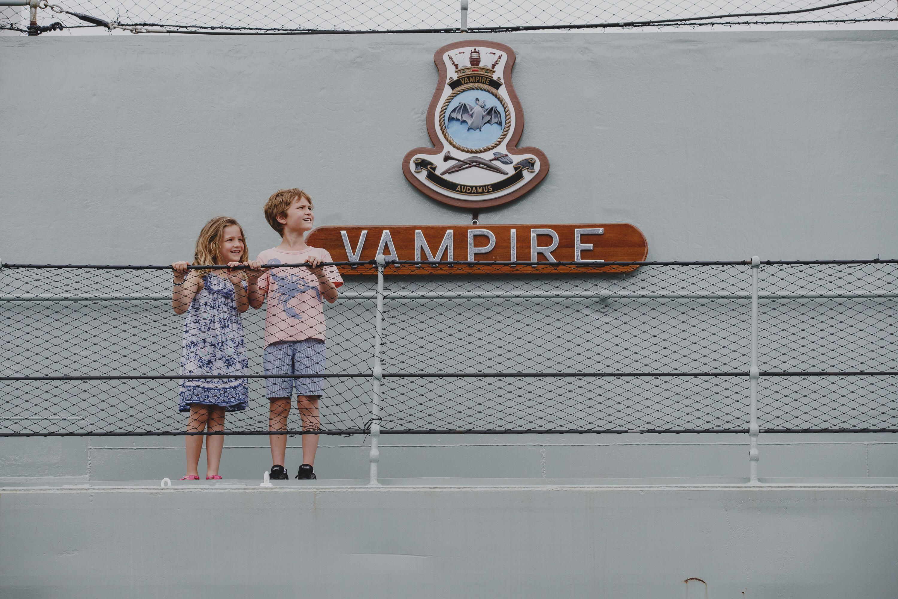 A boy and girl, sister and brother, pose on the upper deck of a navy ship beside the ship's emblem and name Vampire.