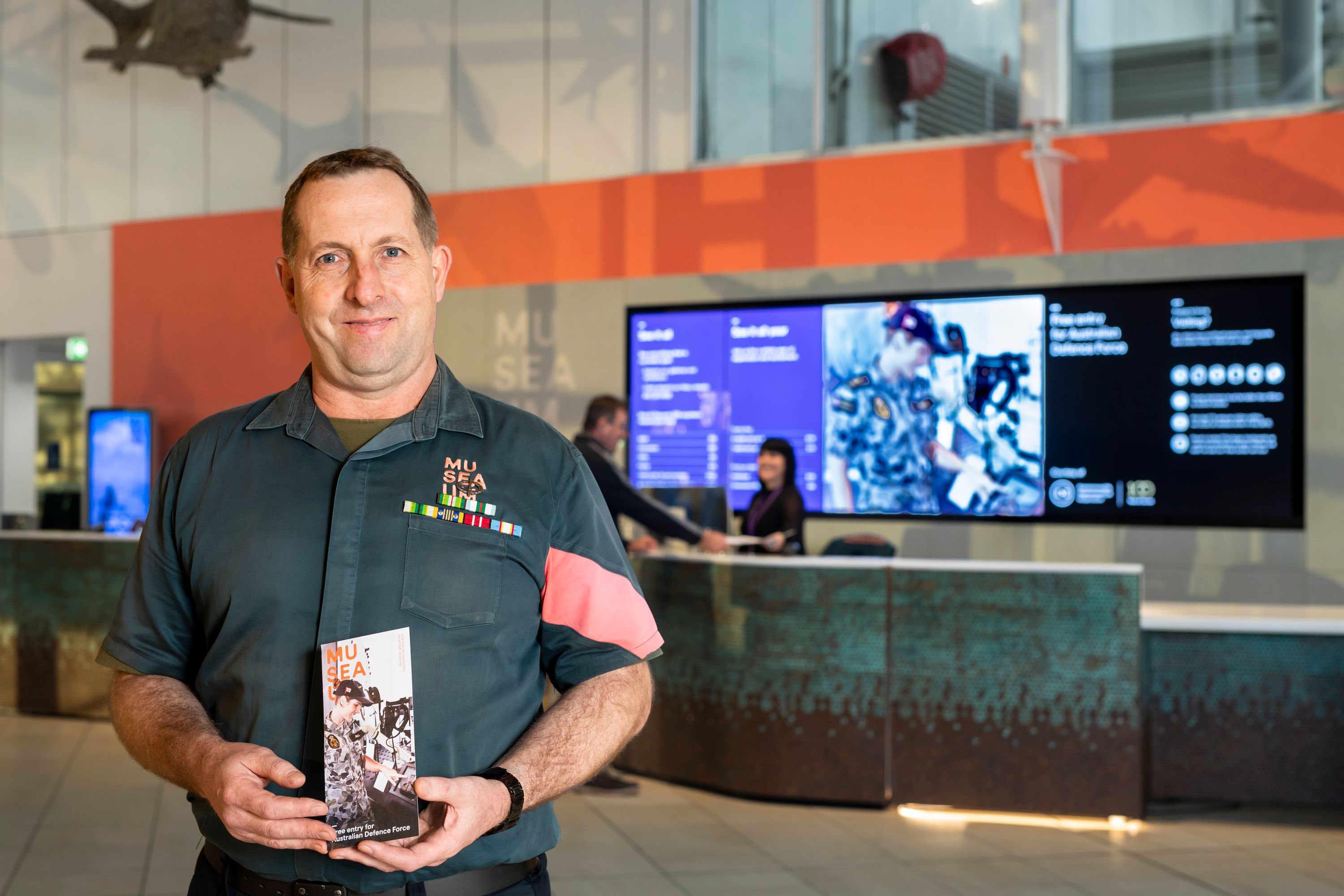 Museum Volunteer in the museum foyer with front of house desk in the background. He holds the museum's 'Free Entry for Australian Defence Force' information brochure.
