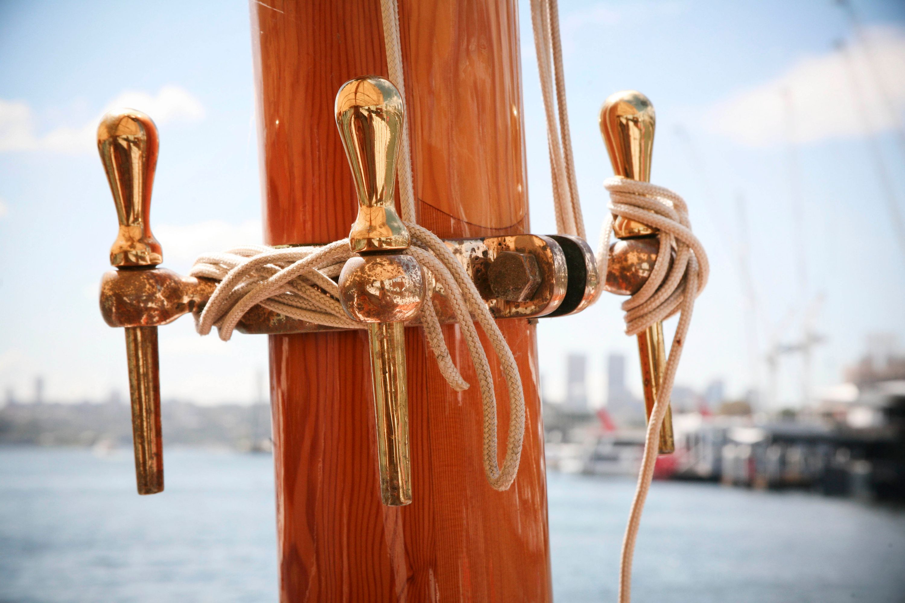 Photograph showing a close up of ropes and brass fittings around a wooden pillar on a boat. The harbour is out of focus behind it.