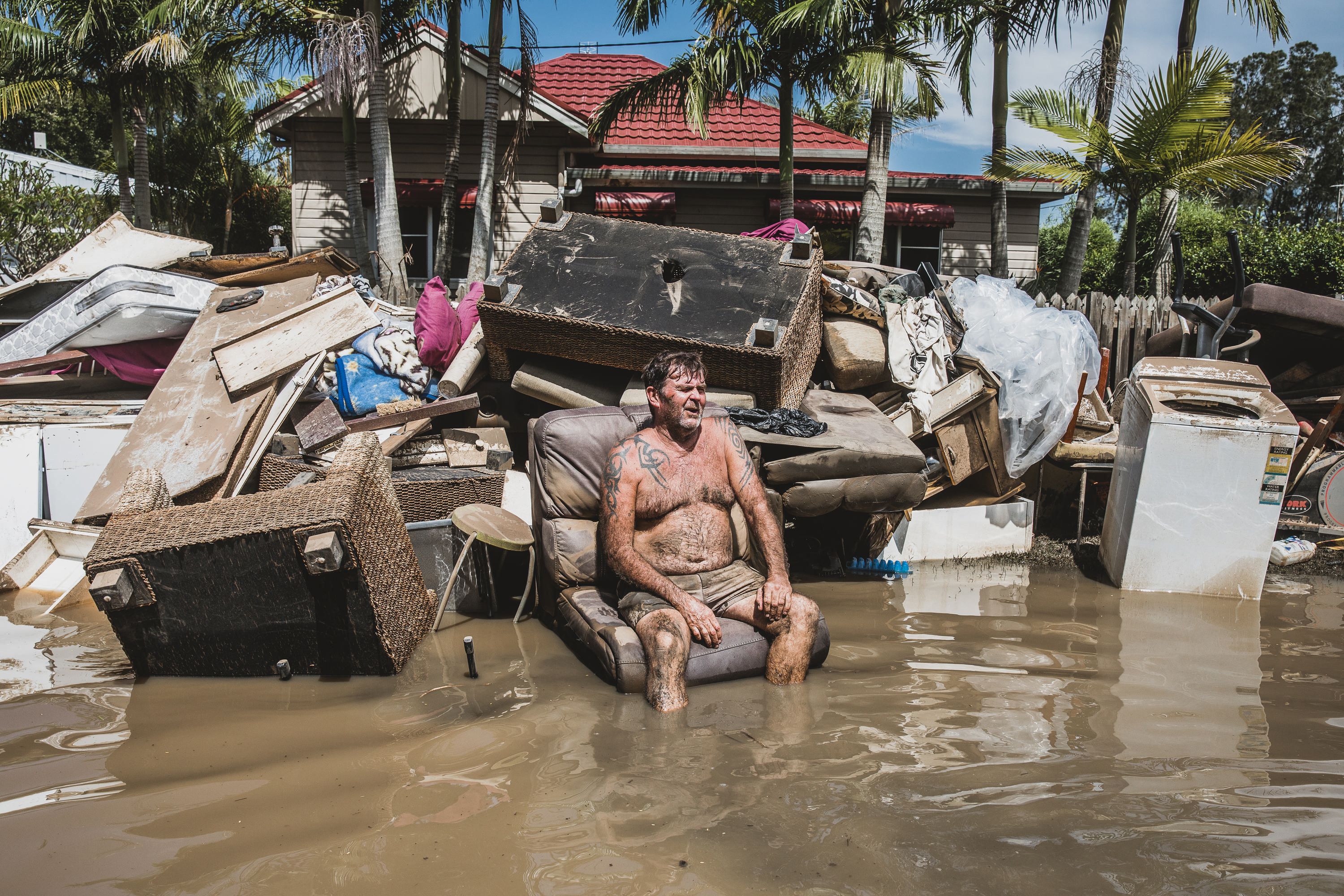 Photo of a man sitting outside, on a seat, surrounded by floodwater, with a pile of destroyed furniture behind him. 