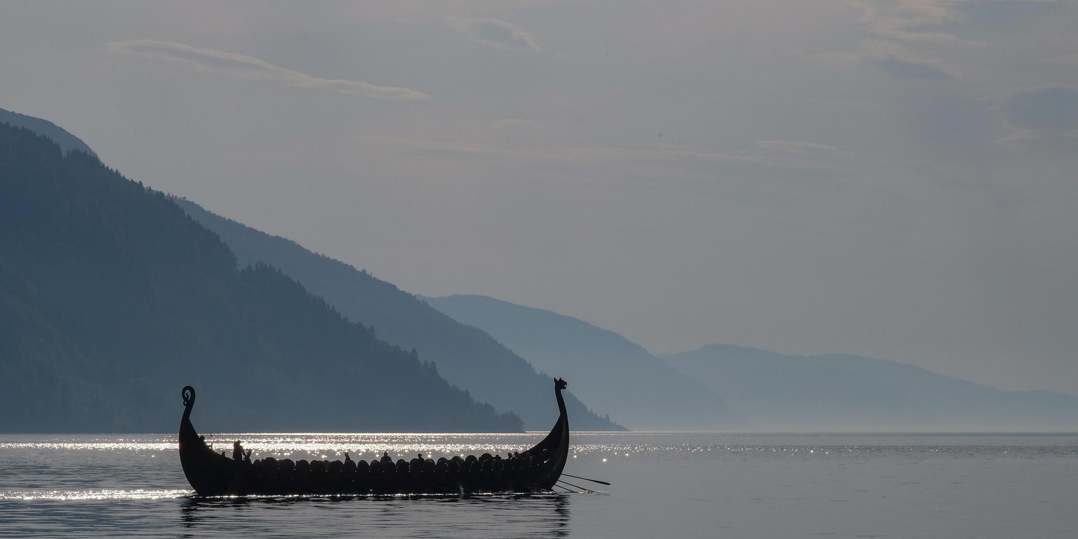 Photo of a wooden long ship on still water with mountains in the background. 