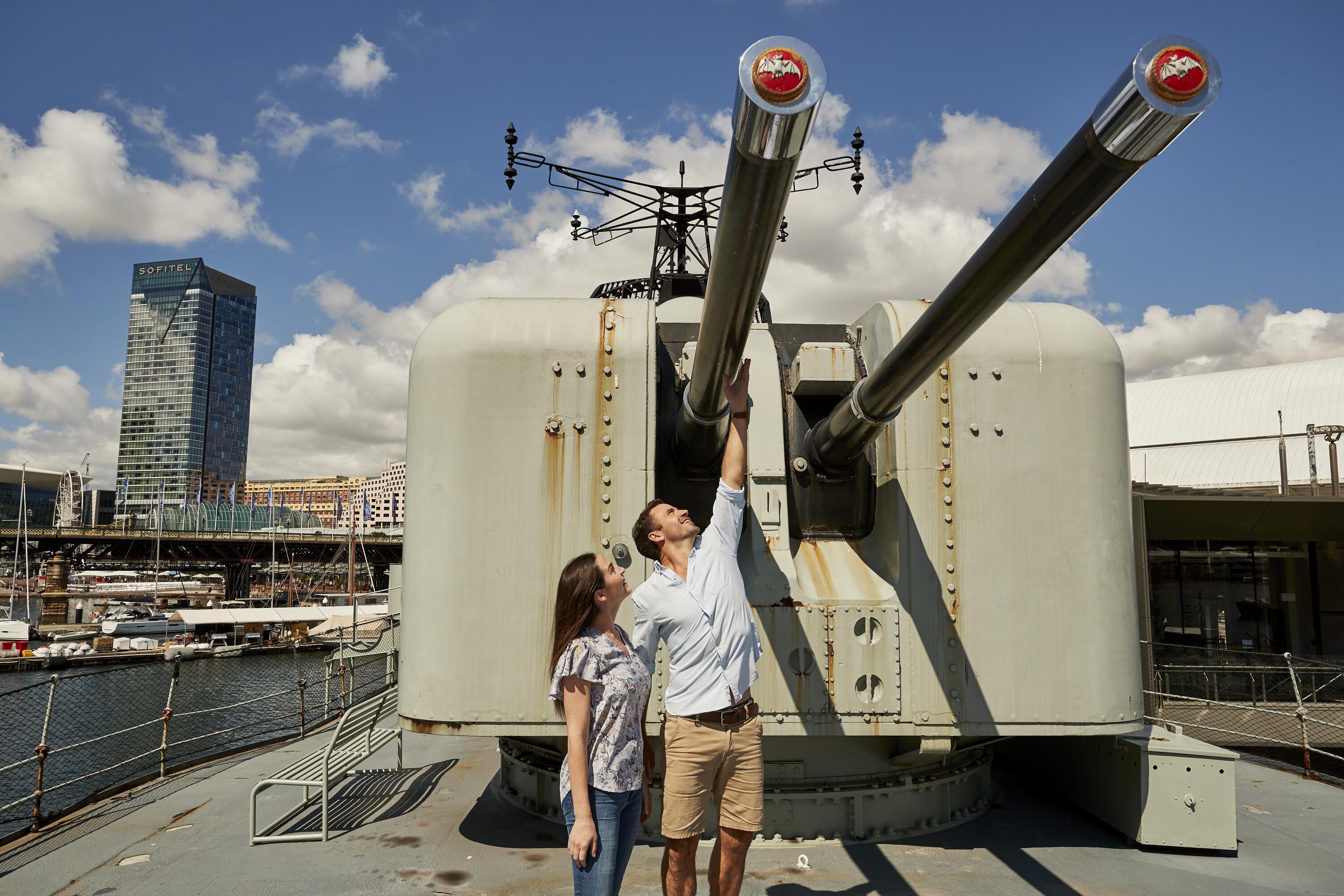 Photo of a couple looking up at the guns on the deck of a Navy warship.