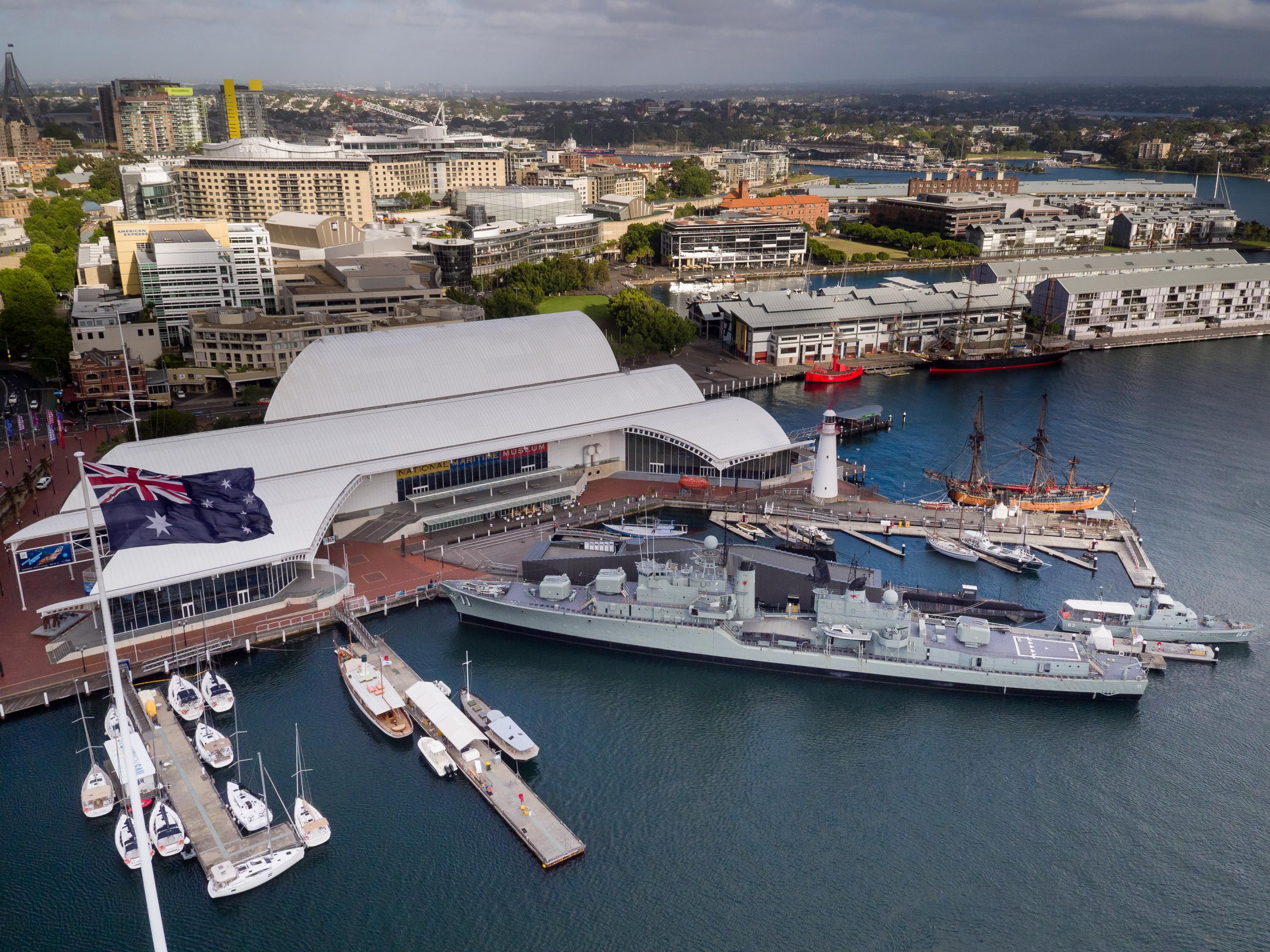 Aerial view of the museum shot by drone. Museum building at centre with Wharf 7 building at upper right. There is an Australian flag, and various wharves and museum vessels.