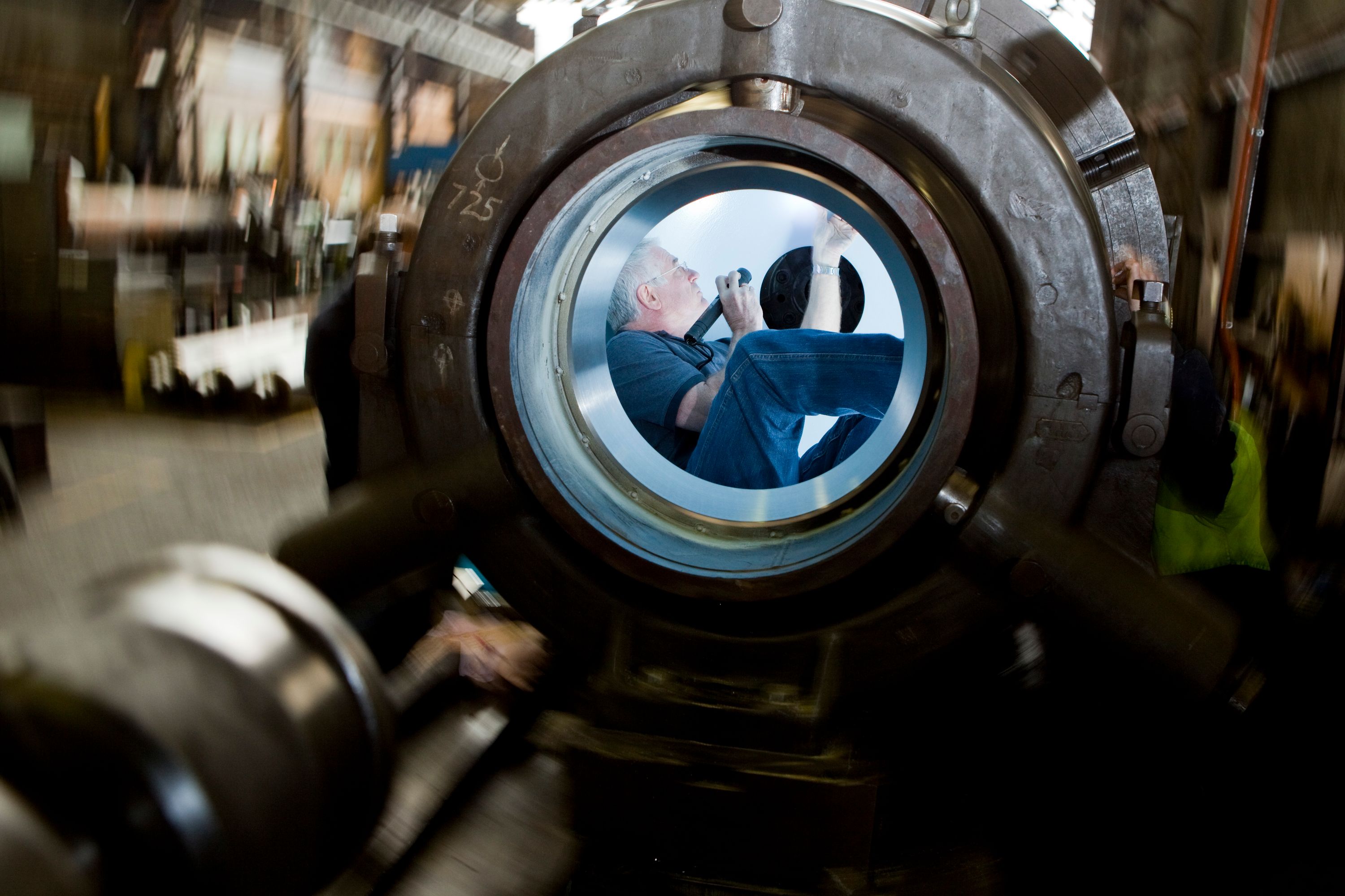 Photo of a man with white hair siting inside a metal sphere, wearing a dark grey shirt and blue jeans. He is holding a torch and examining the inside of the sphere.  