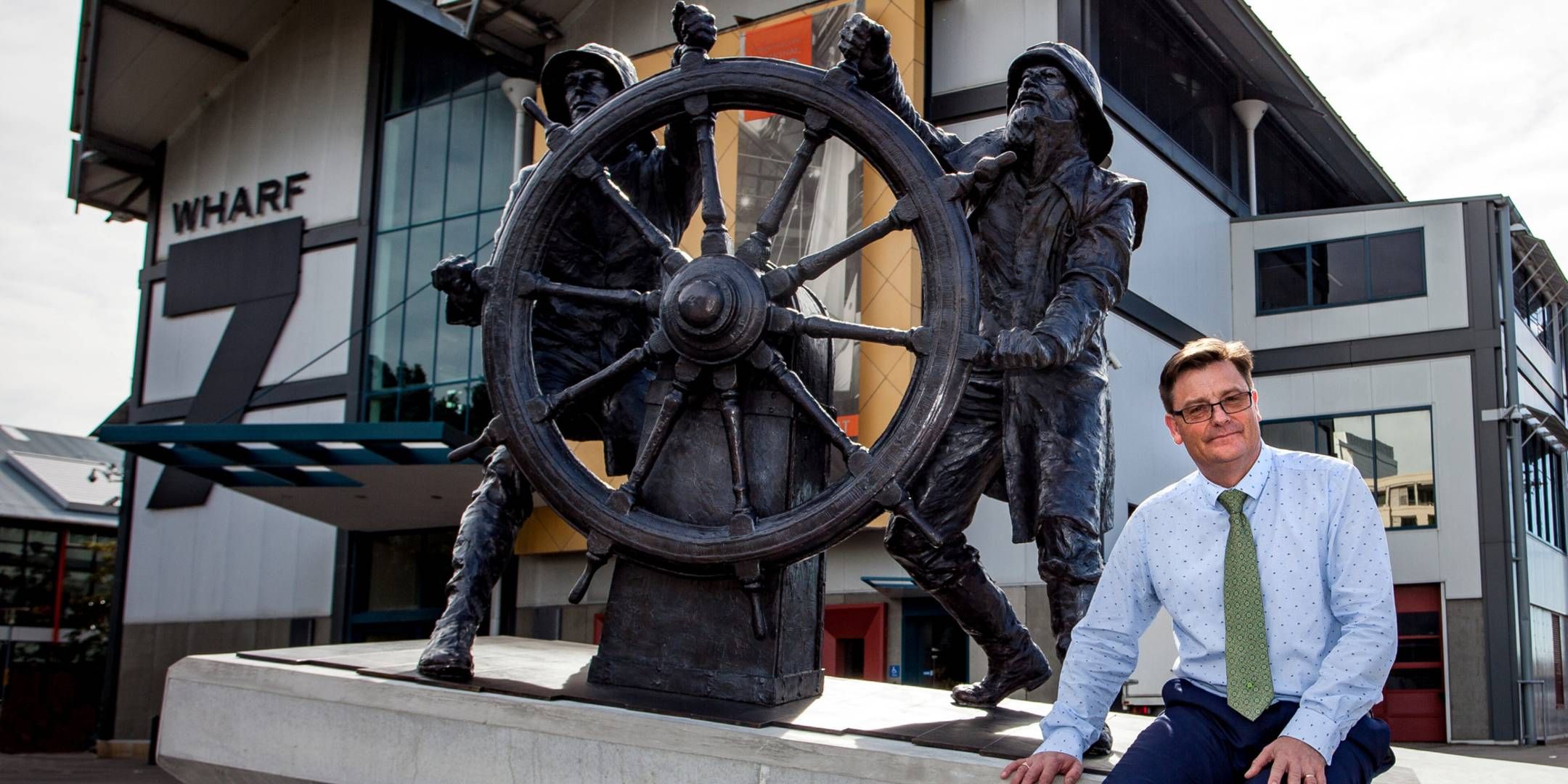 Photo of a main in a dress shirt and tie, seated outside next to a sculpture of sailors.