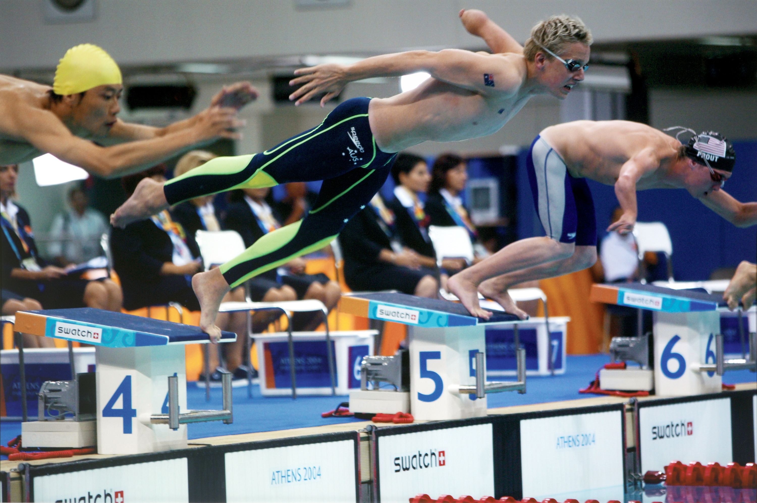 Photo showing 3, male Paralympic athletes diving off the blocks at the stat of a swimming race. 