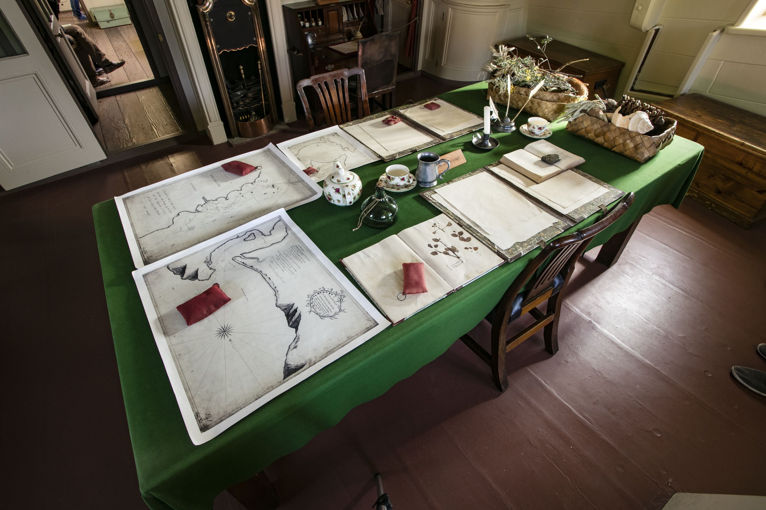Photo showing a table with green cloth and lots of maps and documents arranged on it. There are also 2 baskets of dried, Australian native plants. 