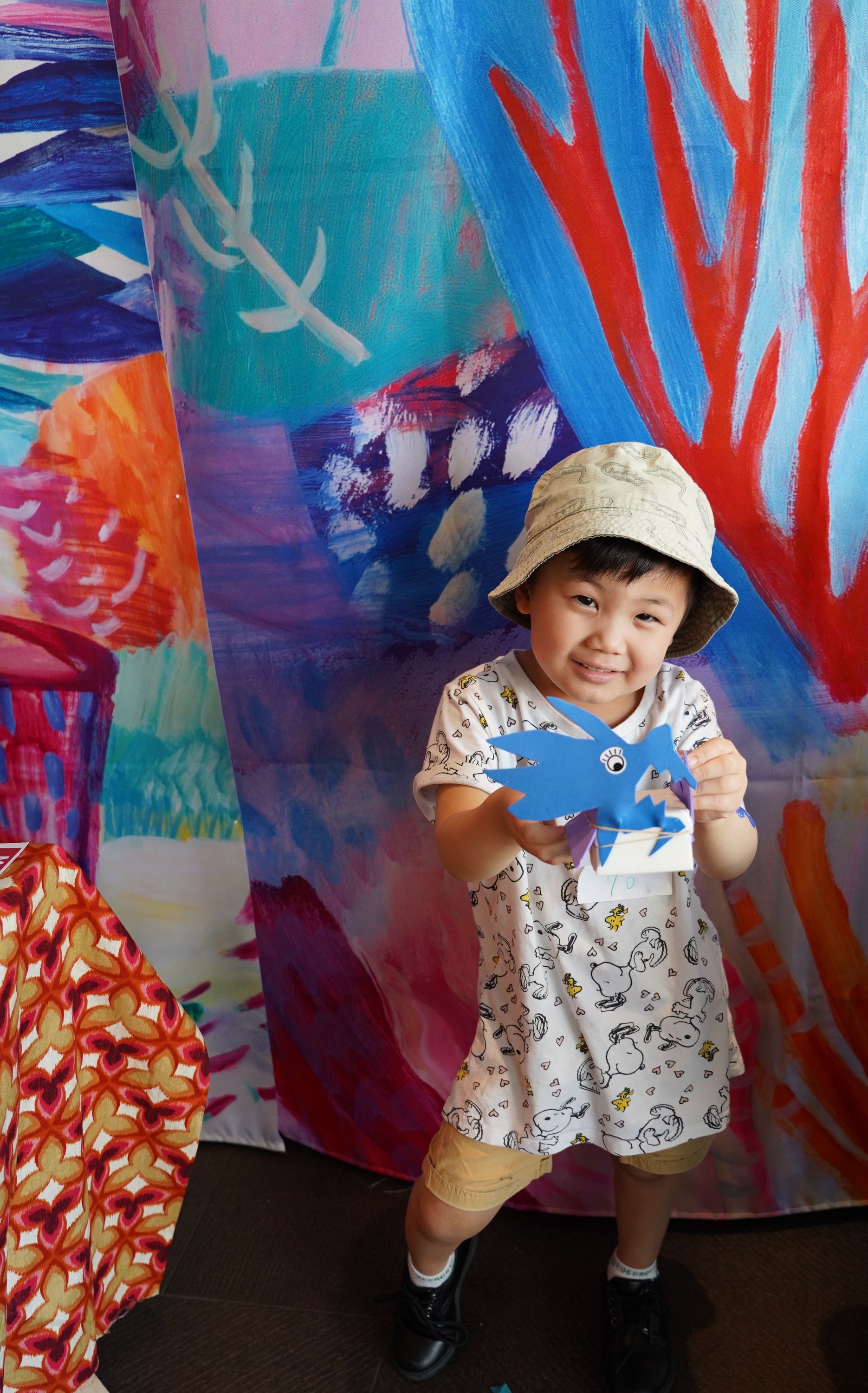 Photo of a small child wearing a cream hat and outfit, holding up a blue dragon head made from paper.