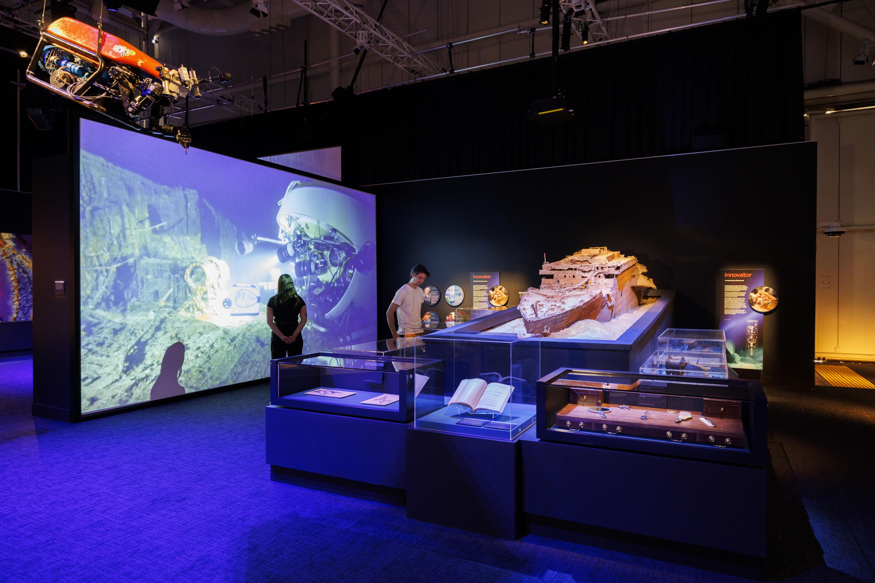 A man and a woman in a museum exhibition. To the left the woman watches a large screen with a robot doing underwater maritime archaeology, while on the right, the man looks at a large ship model and objects in display cases.. 