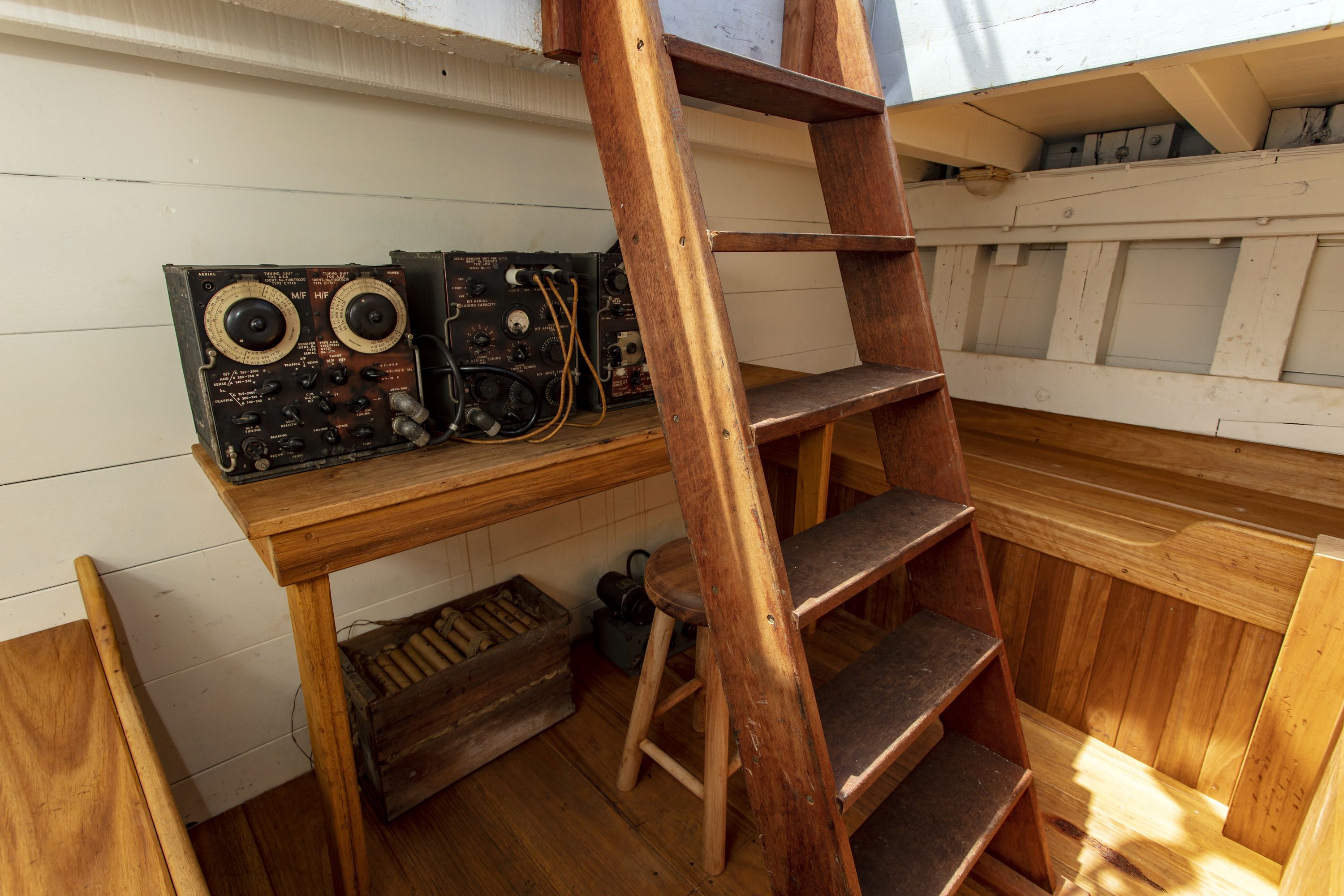 Photo of a small, wooden room below decks on a boat showing radio communications equipment and a ladder. 