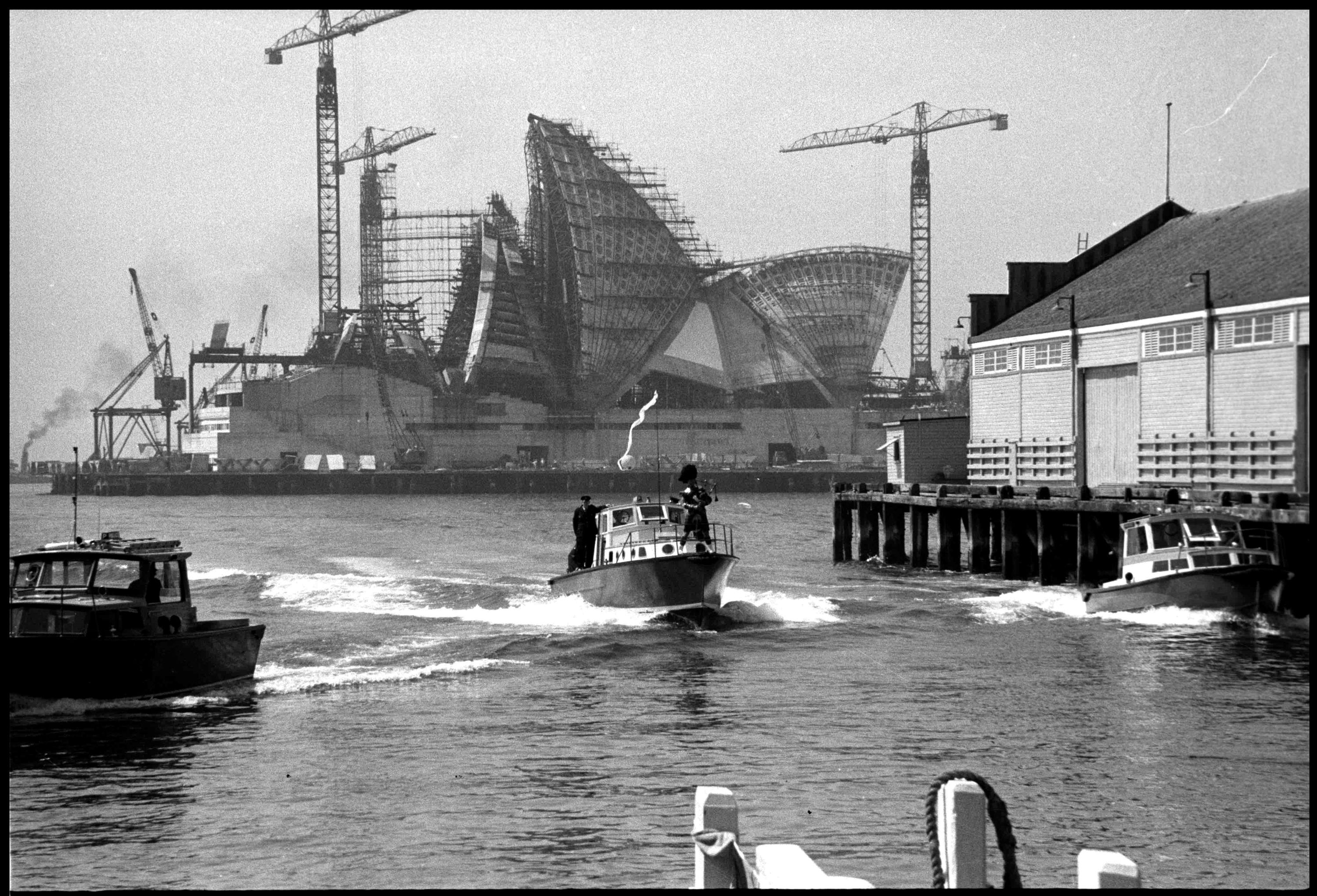 Black and white photograph with a small boat motoring towards the camera with a figure in dark clothing on either side,  the one on the right is playing bagpipes. In the background, the Sydney opera houe is being constructed, with scaffolding and cranes around the part contructed sails.