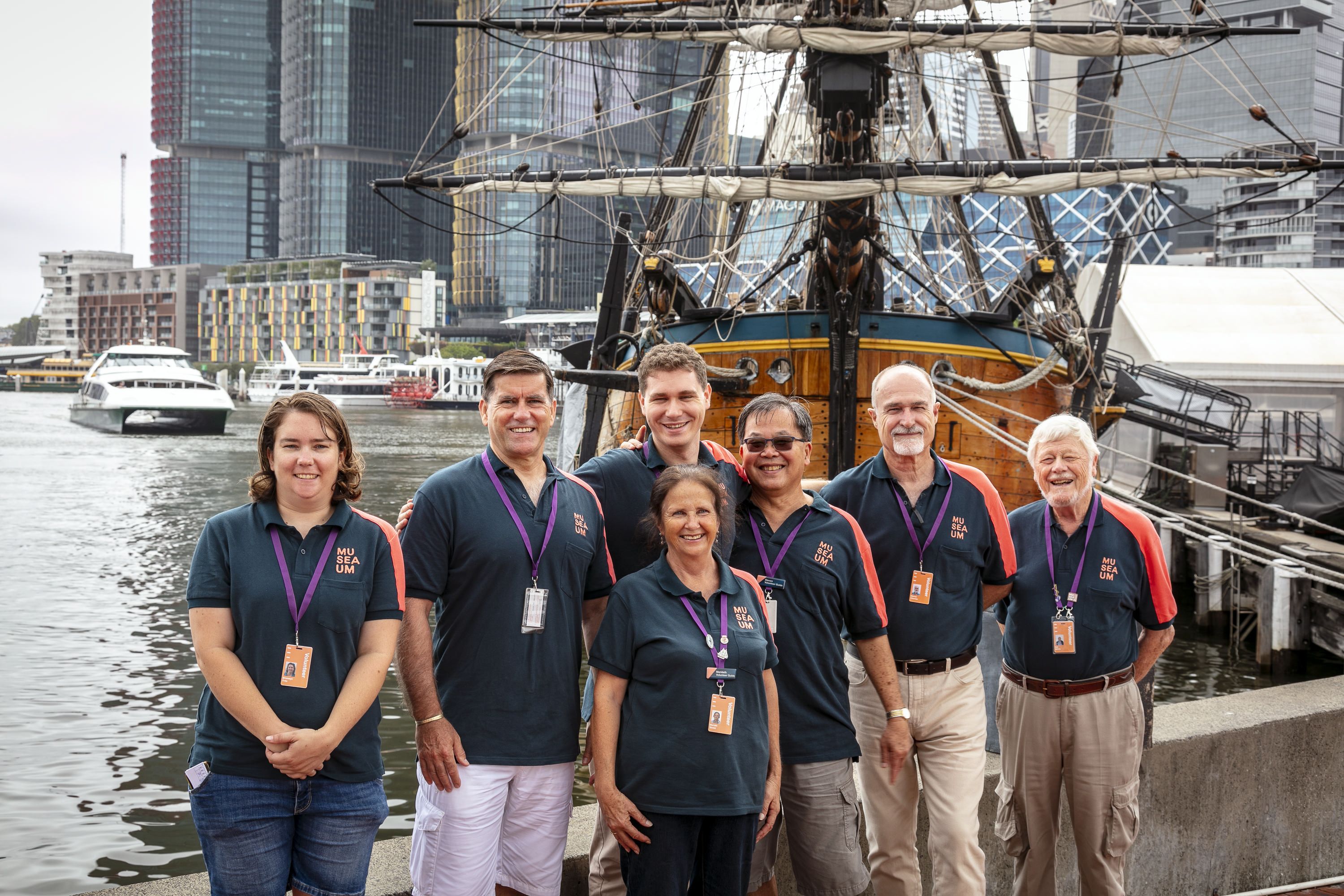 6 volunteers in navy museum unforms posing with HMB ENDEAVOUR replica tall ship in background.