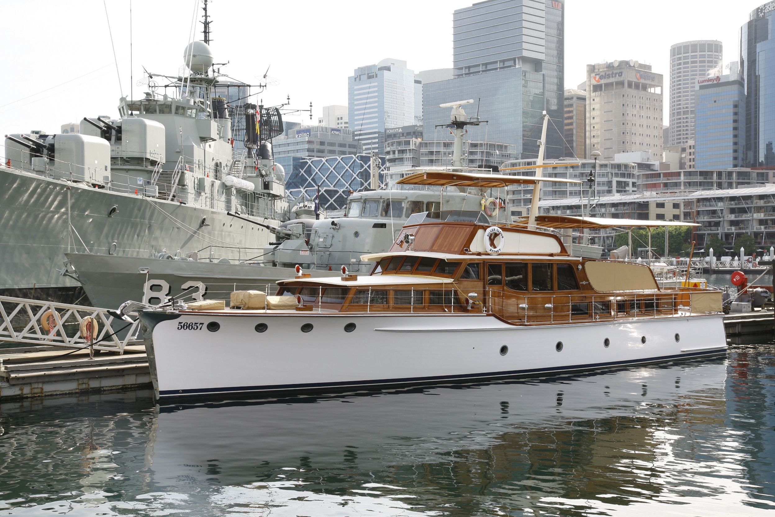 Photograph showing a wooden boat with white hull at the museum wharf, with a navy ship behind it. 