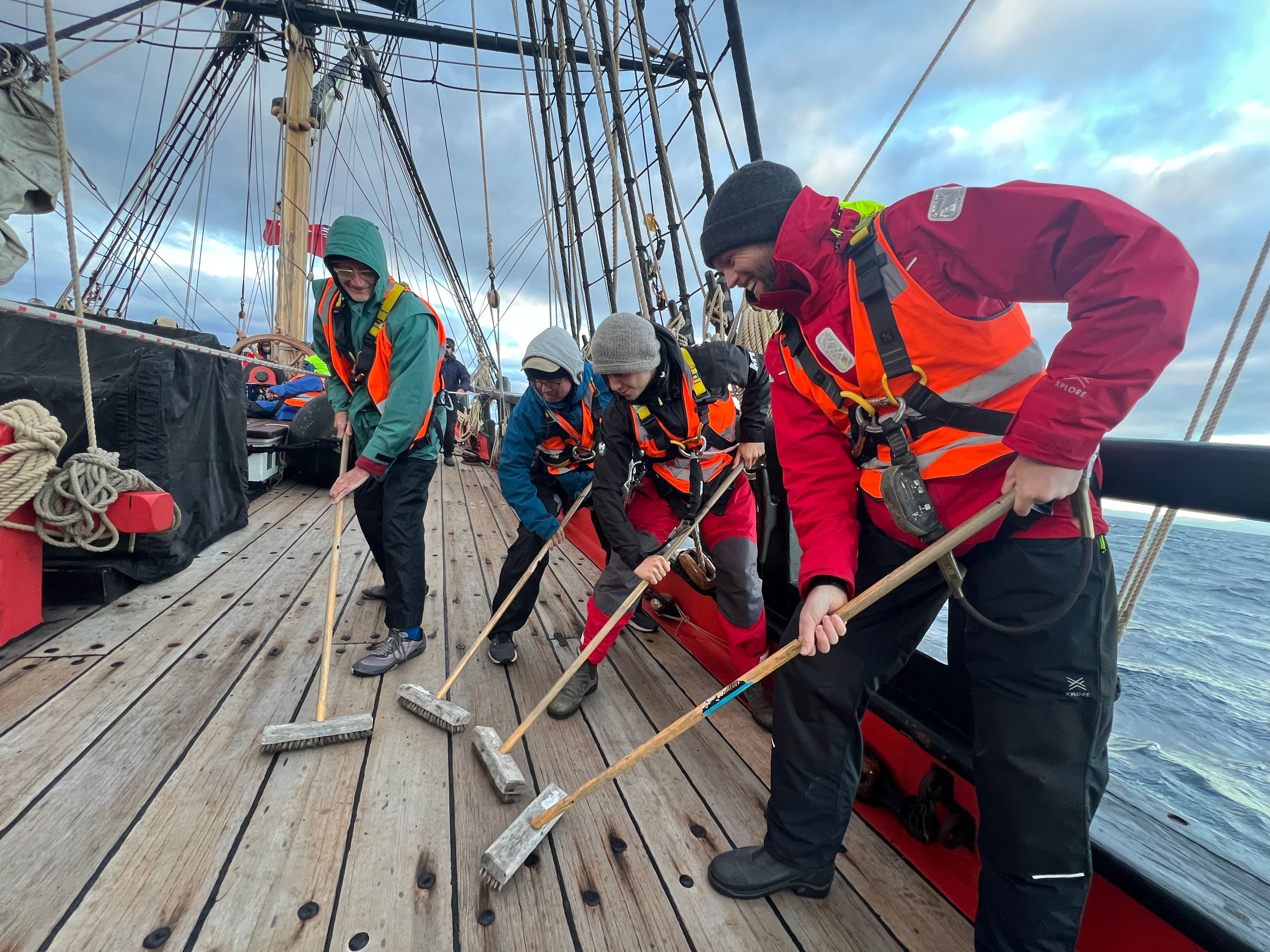 A group of people wearing wet weather jackets and harnesses using brooms to scrub the deck of a tall ship.