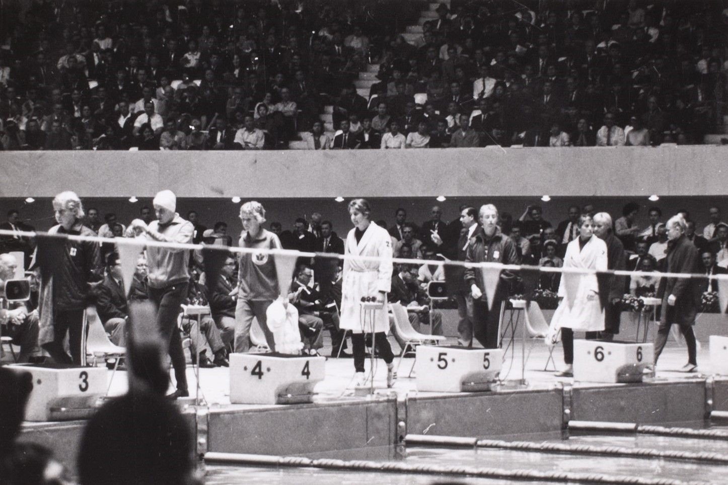 Black and White photo showing a swimmers walking out to one end of a pool preparing for a race with a crown behind them.