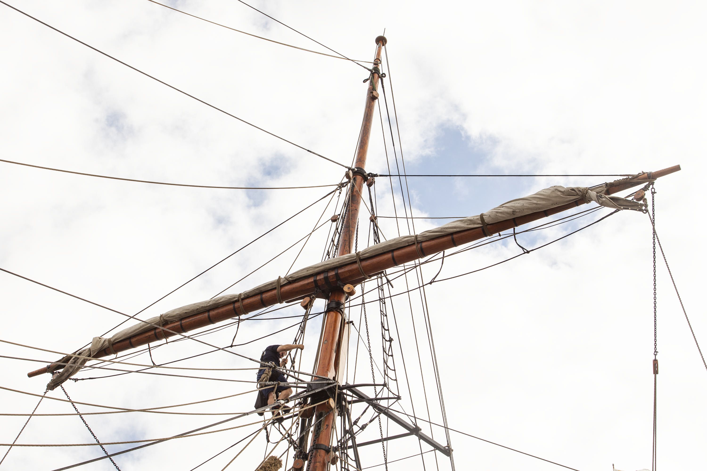 Photo taken looking up at a ship's mast with a person climbing up and white clouds behind. 