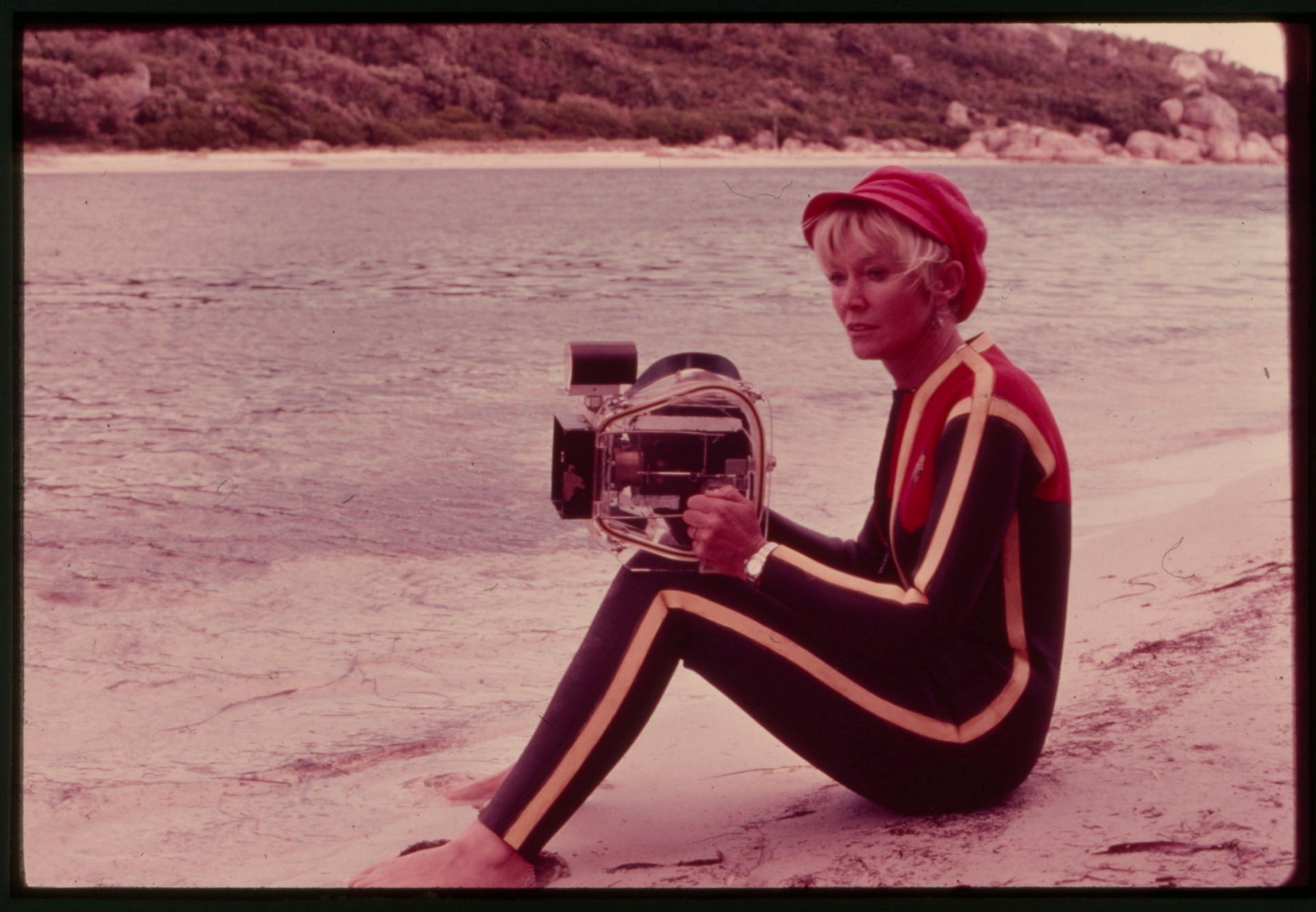 Photograph showing a woman wearing a wetsuit and red hat, siting on a beach and holding a large camera in a protective case.