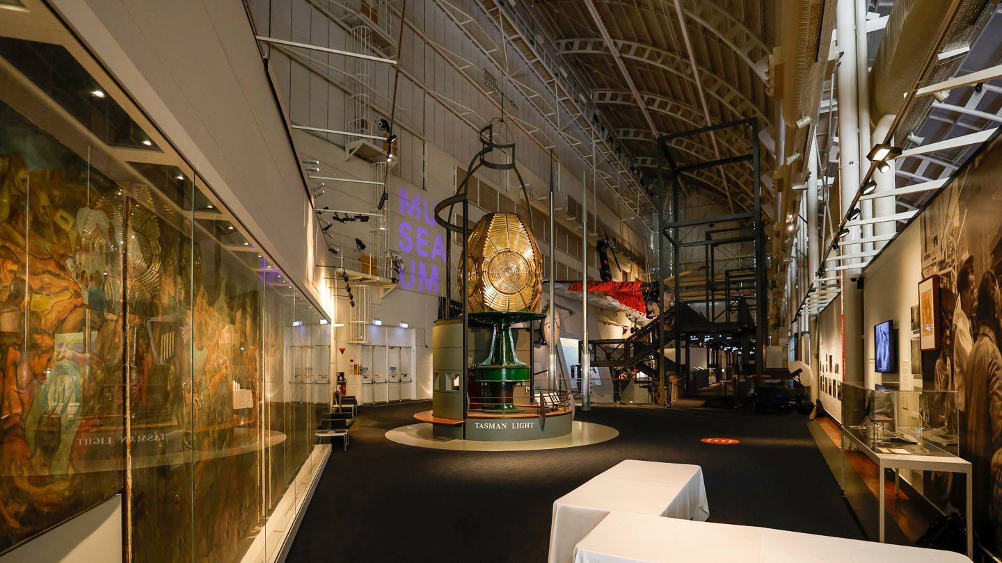 Photo of inside a museum gallery. The gallery features a large lighthouse lantern in the middle of the room and paintings around the sides.