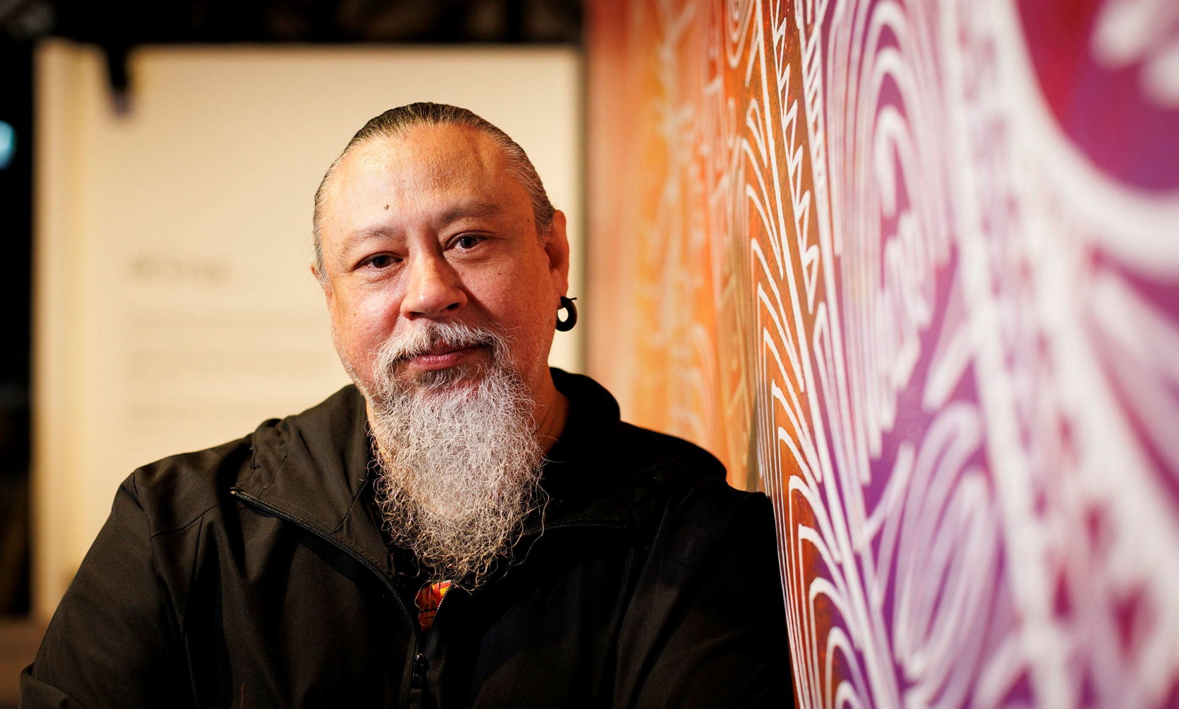 Photo of a man wearing black, with a grey beard and black hair, leaning against a wall featuring a colourful mural.