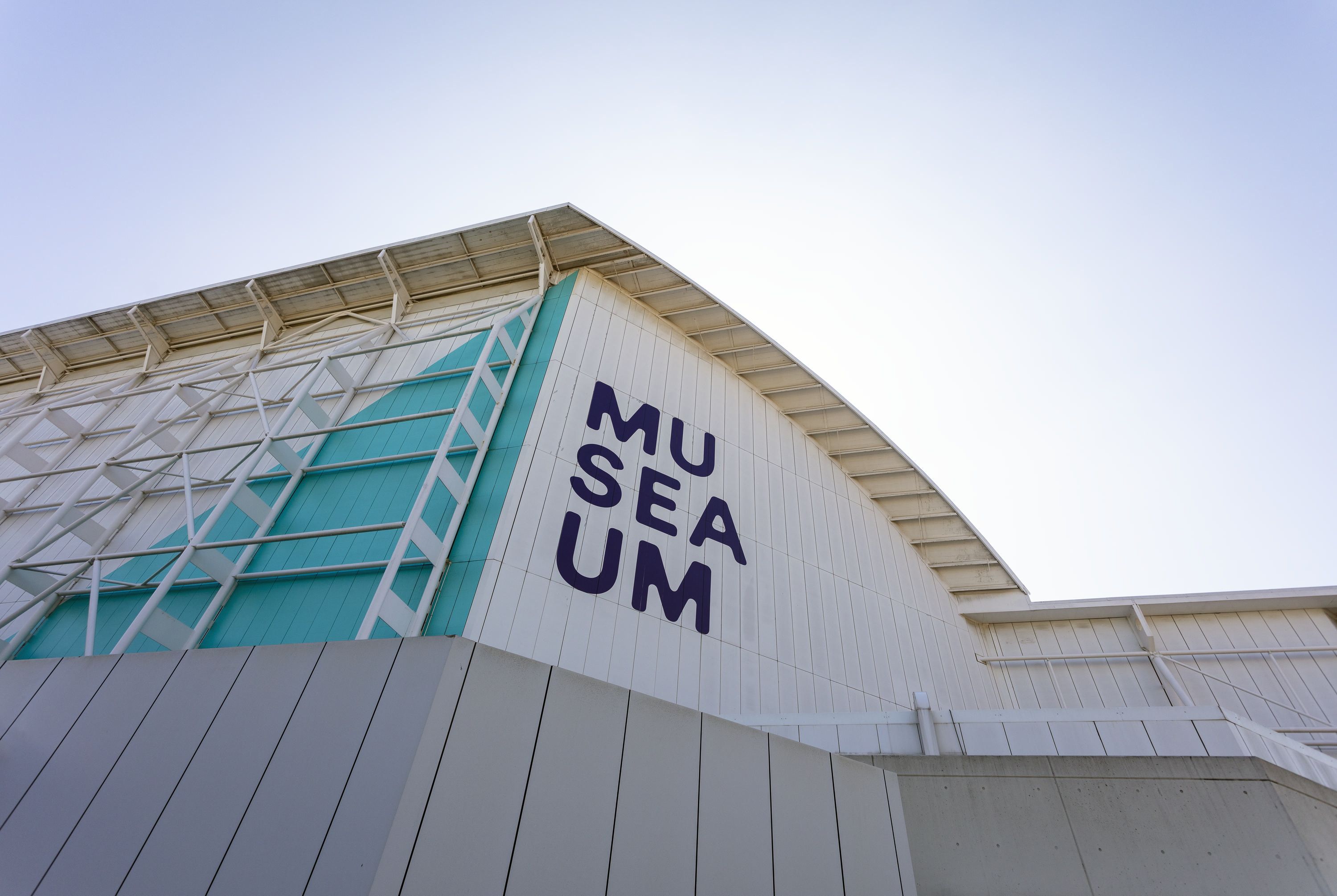 Photo of the museum building, with the logo on the white wall, and a blue sky behind. 