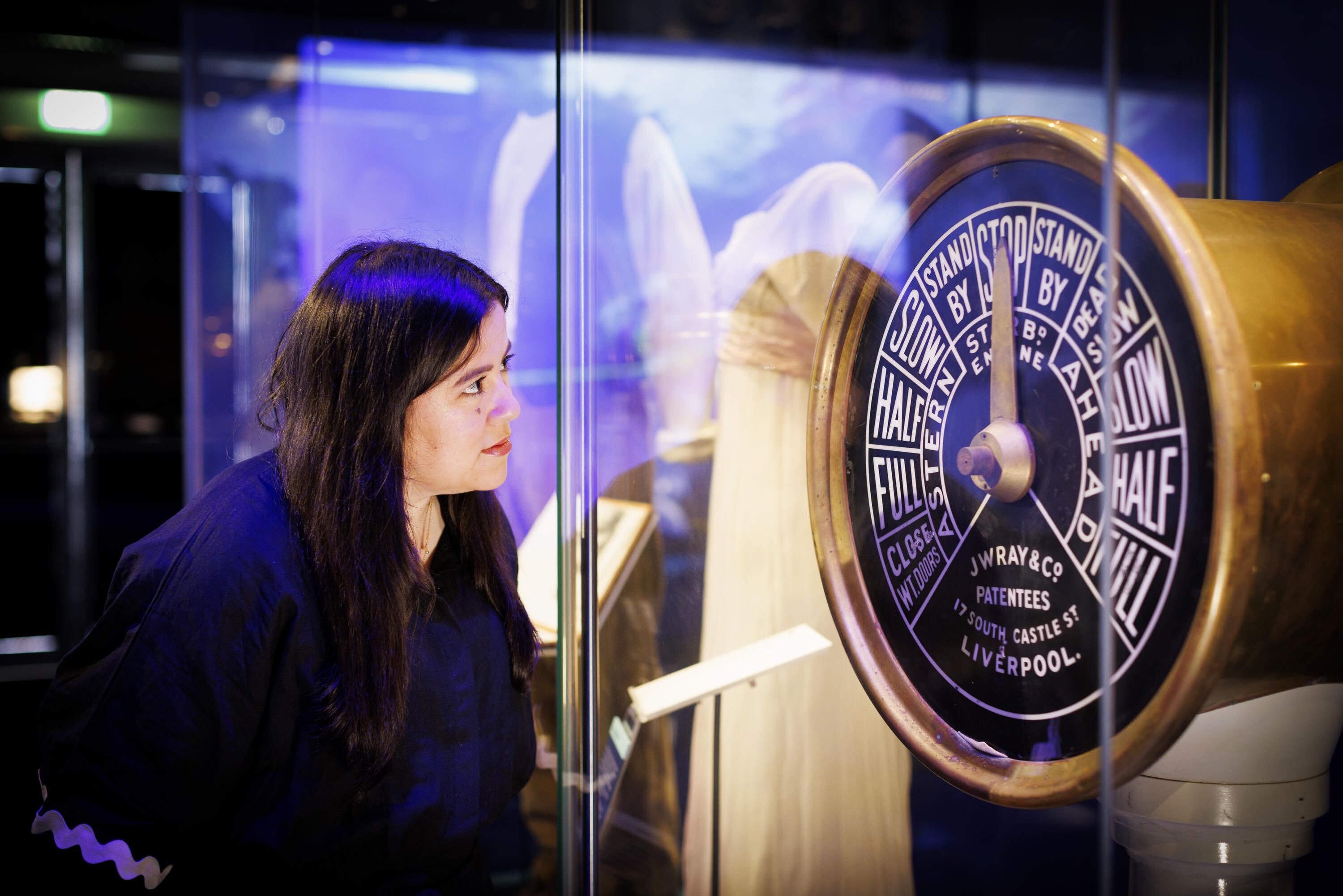 A woman with dark har and clothes looks at a ship's telegraph in a museum display case. 