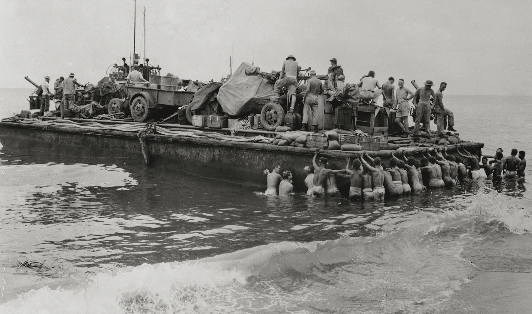 Black and white historical photograph of a barge laden with supplies. There are men on the barge and men in the water.
