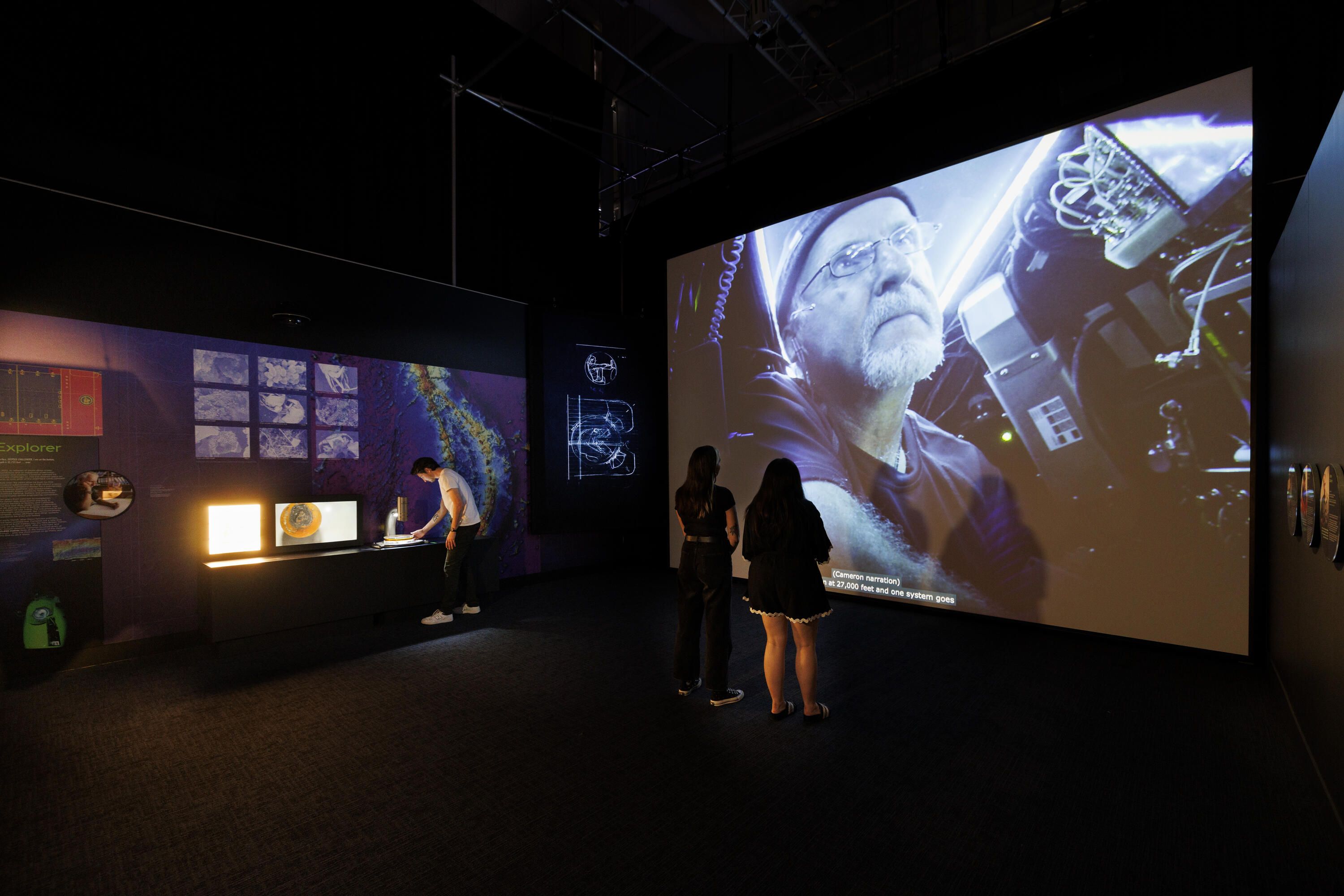 3 figures in a dimly lit museum exhibition. 2 women are watching a large screen projection and on the left of them, a man is looking at a microscope with a screen.  